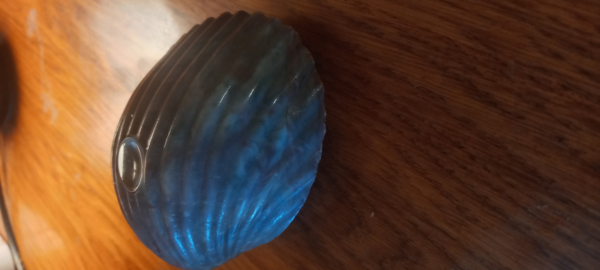 A blue, ribbed object resembling a shell is placed on a brown wooden surface. A clear, circular droplet of liquid is visible on the top center of the blue object. The background is out of focus and appears to be a lighter brown wooden surface. There is no indication of a specific location within the city of Kryvyi Rih, Ukraine.