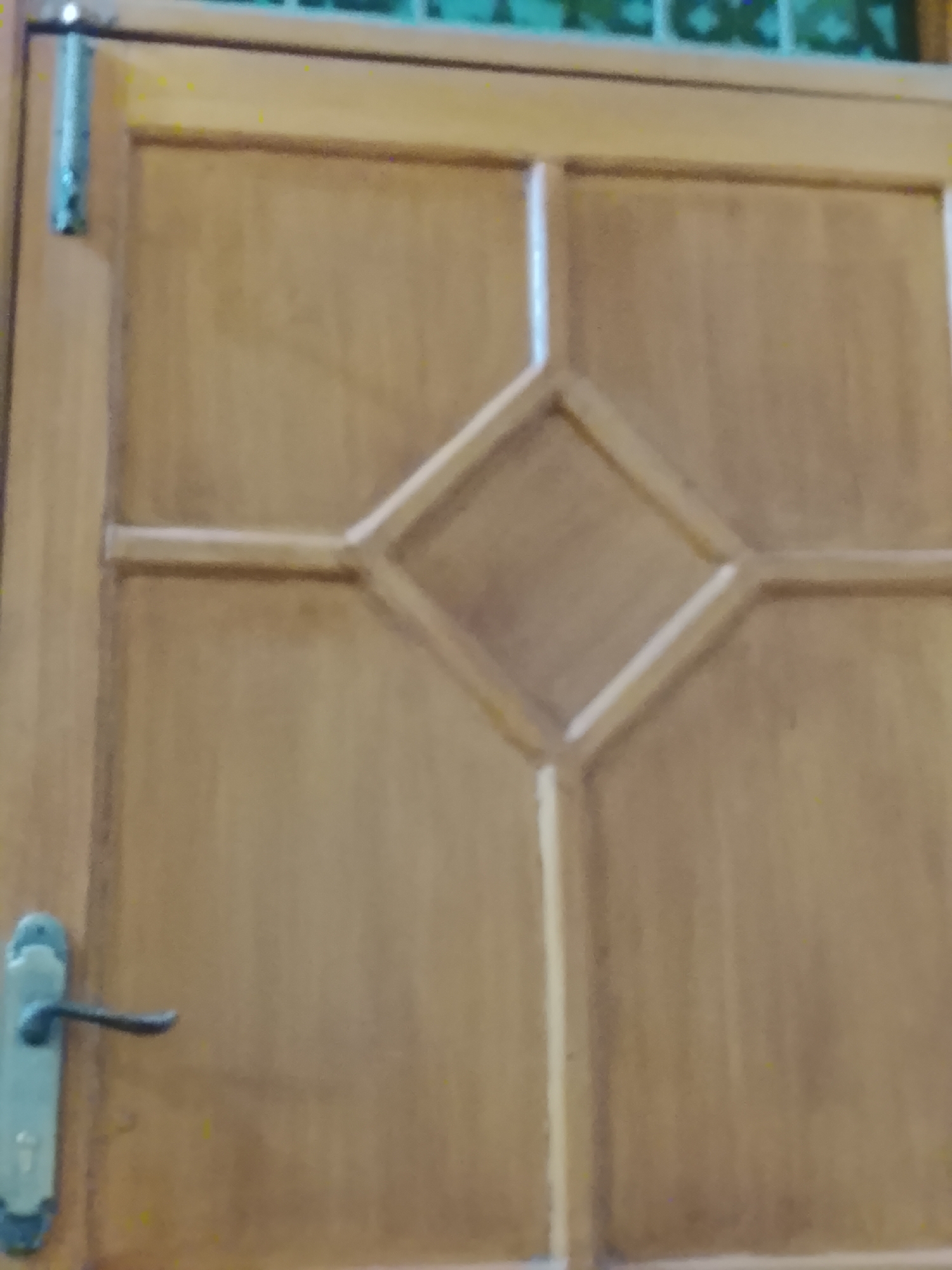 The image shows a light brown wooden door with a diamond-shaped design in the center. A metal door handle is on the left side, and a metal hinge is visible at the top left. The location is Multan, Pakistan, but the specific details about the environment are not discernible from the image.