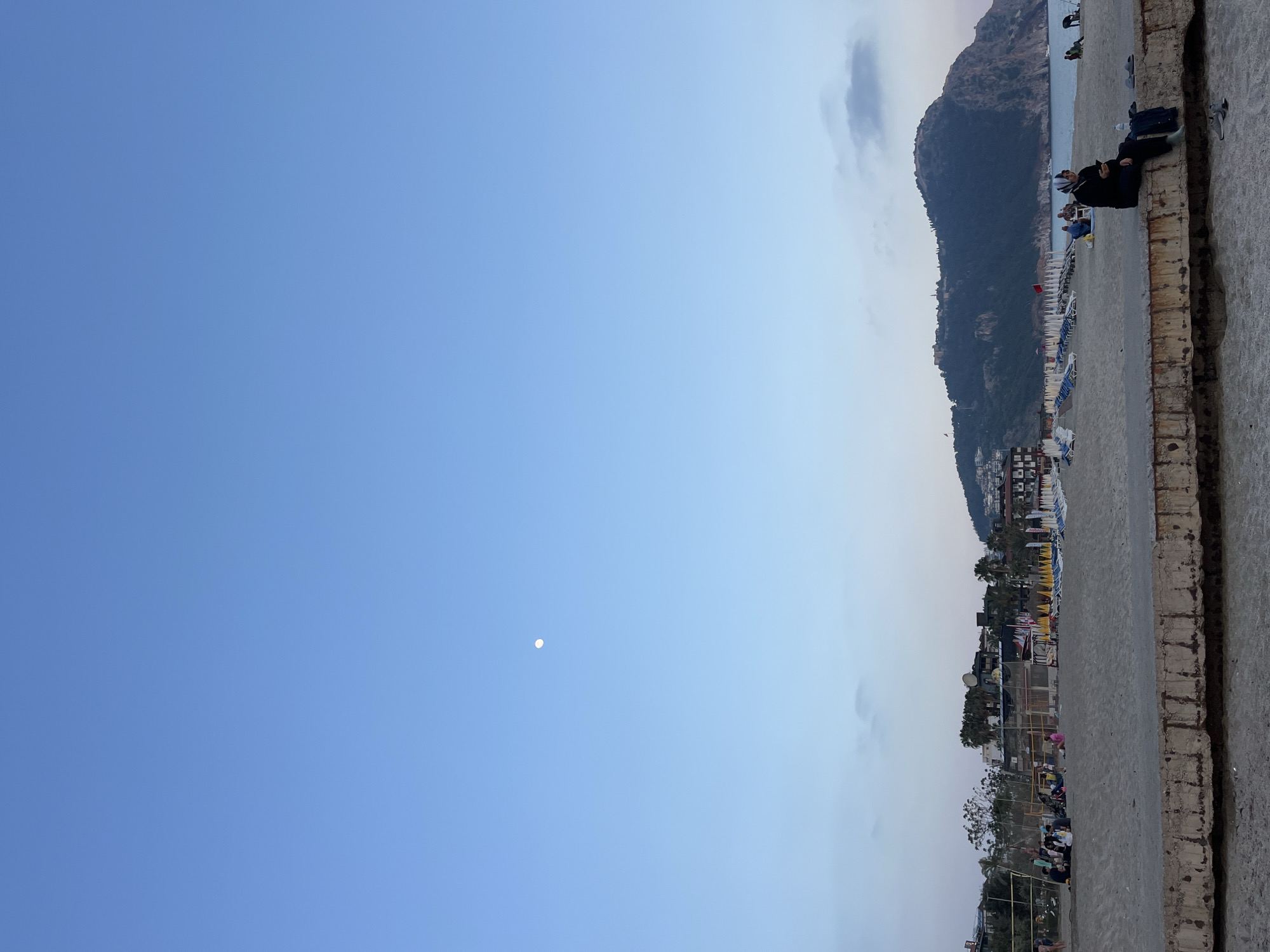The image depicts a serene late afternoon or early evening scene on a beach in Alanya, Turkey. The sky, a clear and soft blue, occupies a significant portion of the frame, with a faint, luminous moon visible towards the center-left.
On the right side of the image, a large, dark green mountain range rises steeply, its peaks partially obscured by a few wispy clouds. At the base of the mountains, a long sandy beach stretches out, populated with numerous beach chairs and sun umbrellas, many featuring yellow, blue, and white stripes. Along the coastline, various buildings, including hotels and resorts, dot the landscape, showcasing a developed coastal town. The calm, blue sea is visible beyond the beach, with what appear to be boats in the distance.
In the foreground, a weathered concrete wall or barrier runs parallel to the beach. Several people are scattered across the beach and seated on this concrete structure. One individual, dressed in dark attire, is clearly visible seated on the wall, seemingly engrossed with a mobile device. Other people are relaxing in groups on the sand, some near the water's edge, enjoying the tranquil atmosphere.
The lighting is gentle, indicative of the sun setting or having recently set, casting a soft glow over the scene. The overall impression is one of peaceful leisure at a scenic coastal location. No clear, readable text is visible in the image.