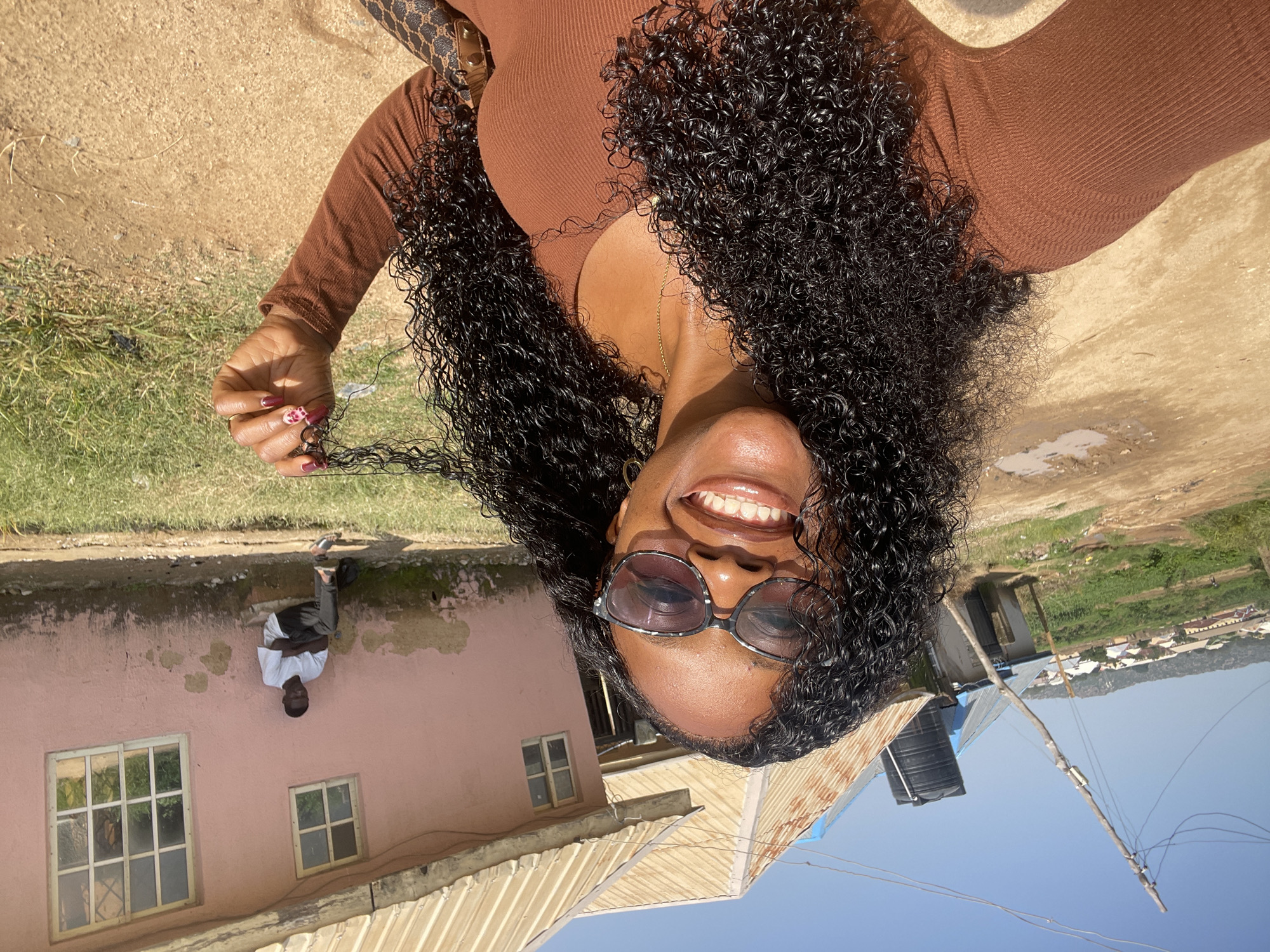A woman with curly black hair and sunglasses smiles while taking a selfie outdoors. She is wearing a brown long-sleeved shirt. Behind her, a man in a white t-shirt and dark pants sits on a step outside a pink building with a corrugated metal roof. The scene appears to be in a rural or semi-rural area, with dirt roads, grass, and a distant view of houses and hills. The location is specified as Rafin Par, Nigeria.