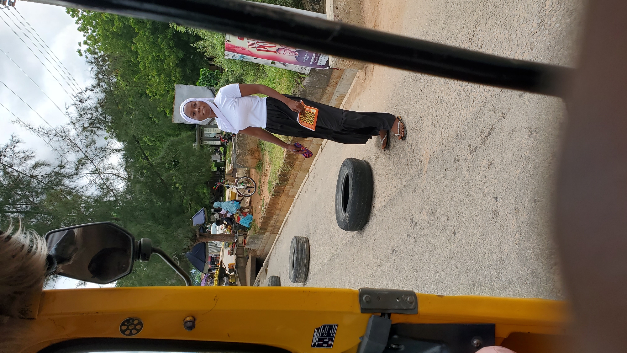 In Jalingo, Nigeria, a woman wearing a white hijab and a white t-shirt, paired with a black skirt, stands on a paved road. She is holding a fan in her right hand. To her left, two used tires are placed on the road. In the background, a yellow vehicle, partially visible on the left, appears to be a tuk-tuk. Further back, there are market stalls with people and umbrellas, a bicycle, and trees lining the street. A poster featuring a man's face is visible on the right side of the frame. The sky is overcast.