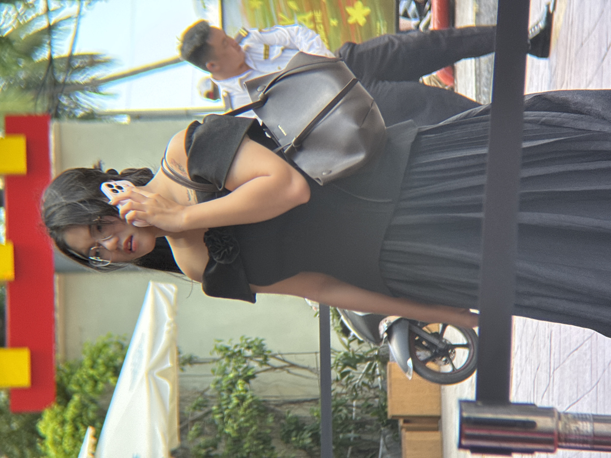 A young woman with dark hair and glasses is on her phone, wearing an off-the-shoulder black dress and carrying a dark brown handbag. She is standing in front of a black pleated skirt. In the background, a man in a white uniform is visible. The scene appears to be outdoors with trees, a red and yellow structure, and a white umbrella. A motorcycle is parked behind the woman. The ground is paved with light-colored tiles. This scene is located at the Nha Trang center, a shopping mall in Nha Trang, Vietnam, where a promotional event is being held.