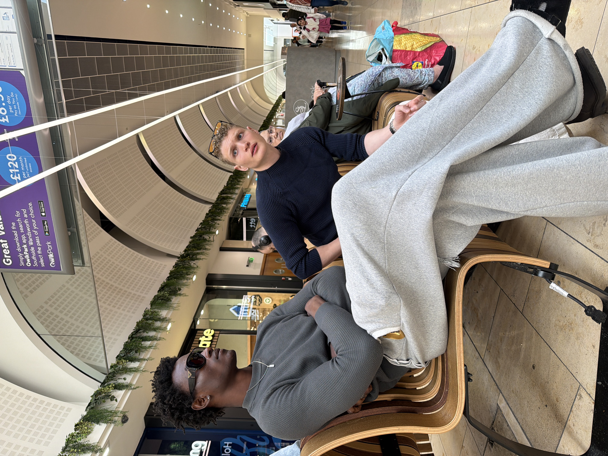 The image captures a scene inside a shopping mall in the city of London, United Kingdom. Three individuals are seated on a modern-looking wooden bench with a metal frame.
A young Black man with a dark curly afro and sunglasses is seated with his arms crossed. He is wearing a grey waffle-knit sweater and a silver chain necklace.
Beside him, a fair-skinned man with short blond hair and glasses is looking toward the camera. He is wearing a dark blue long-sleeved shirt.
Beyond them, another individual is partly visible, seated further down the bench with colorful shopping bags nearby.
The mall's interior features a curved ceiling design with recessed lighting. Several stores are visible in the background, including one labeled "Primark" and another partially visible with the text "Ho". Green garland decorations are mounted above the storefronts. Signage promoting a parking app is posted on the left side of the image, indicating pricing for parking. Several additional individuals are walking in the far background.