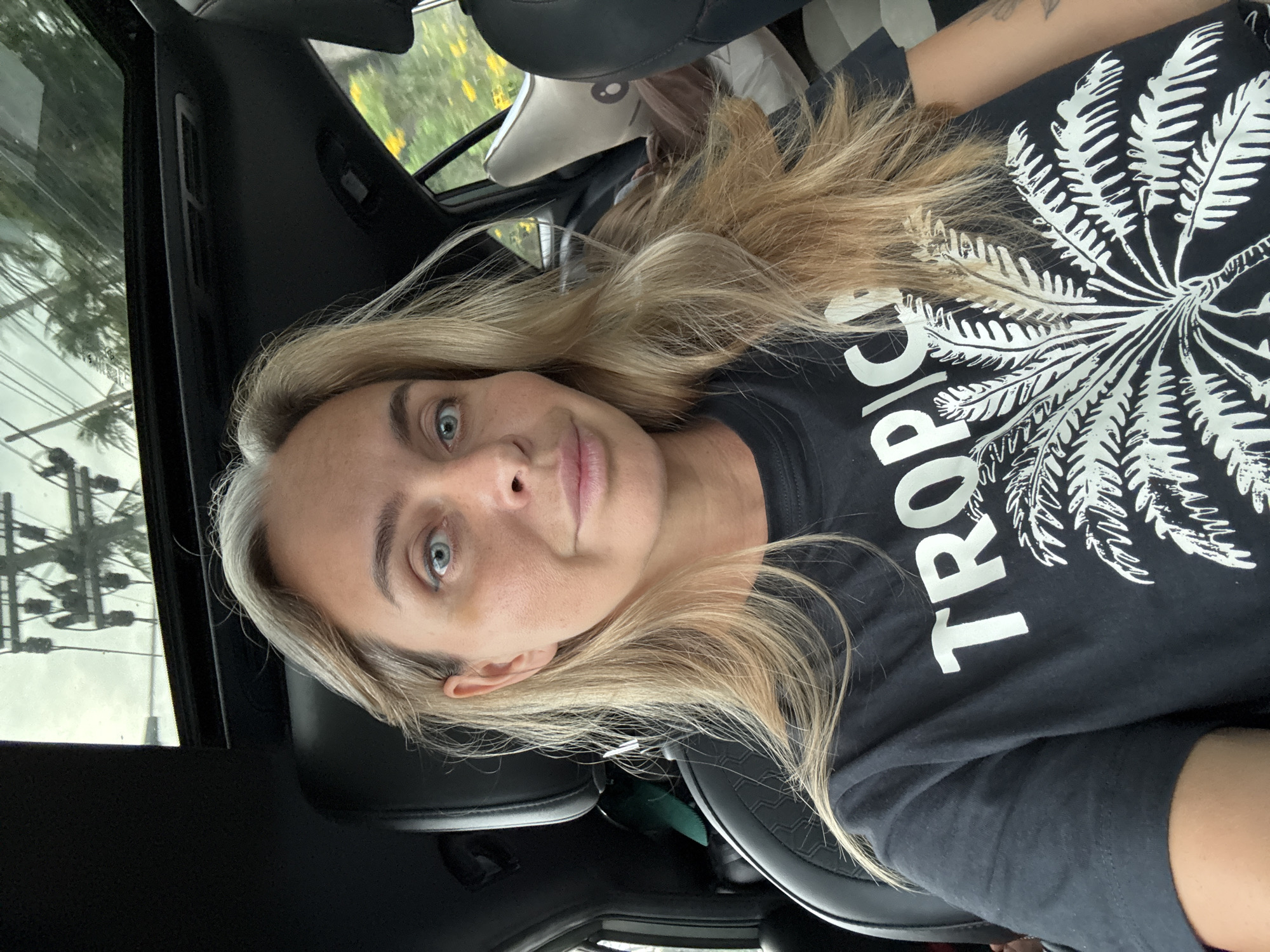A woman with blonde hair and blue eyes, wearing a black t-shirt with a white palm tree graphic and the word "TROPIC" printed on it, is taking a selfie in a car. She is sitting in the driver's seat, and the car's sunroof is open, revealing a cloudy sky and overhead power lines with palm trees in the background. The interior of the car is dark, with black leather seats visible. In the backseat, a child can be seen sleeping, covered by a blanket. The scene appears to be set in Ko Samui District, Thailand, indicated by the presence of palm trees and the general outdoor environment.