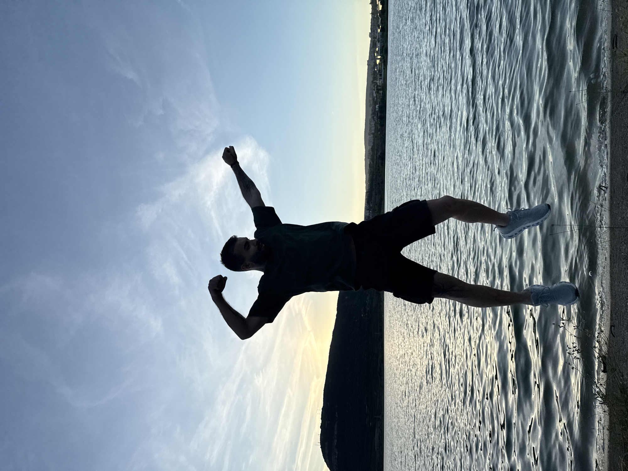 A silhouetted man is captured mid-air, jumping over the water's edge. He is wearing a dark t-shirt, dark shorts, and light-colored athletic shoes. His arms are raised in a triumphant gesture, with his right fist clenched and his left arm extended outwards. The scene is set at dusk with the sun setting behind a dark, hilly silhouette of the landscape. The water reflects the soft, muted colors of the twilight sky. In the background, a city skyline is faintly visible along the horizon. This location is believed to be near the Shumen Plateau in Shumen, Bulgaria.