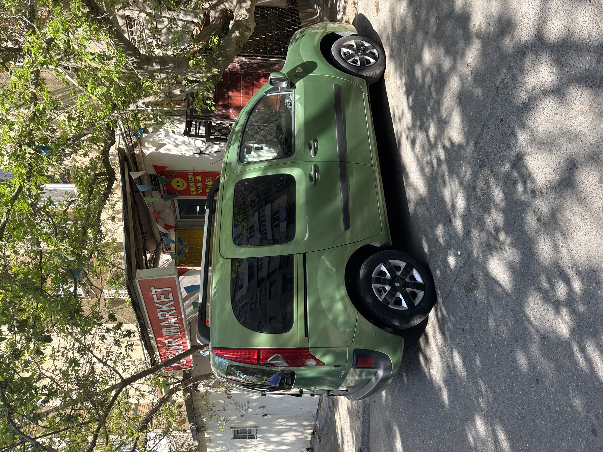 A light green, four-door van is parked on a paved street in front of a small market building. The market has a red sign that reads "UR MARKET" in white letters. There are small, colorful triangular flags strung across the front of the market. The van has tinted windows and a roof rack. Sunlight filters through the leaves of a large tree in the foreground, casting shadows on the street and the van. The background shows a building with windows. The scene appears to be in Baku, Azerbaijan, based on the text on the market sign and the architecture. There are no people visible in the image.