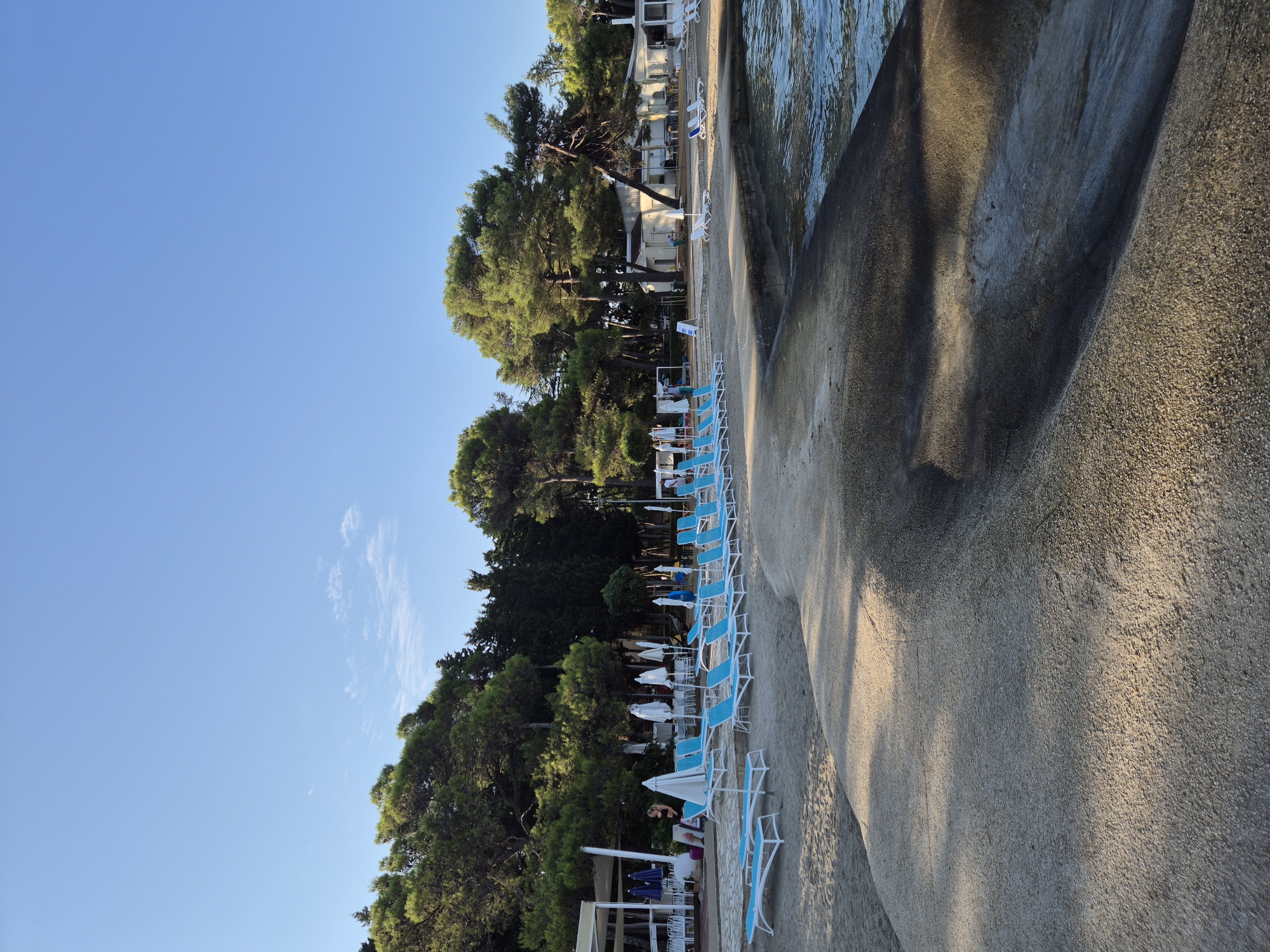 This is a daytime, eye-level shot of a beach area in Monterol, Croatia. The foreground features a smooth, grey concrete walkway or pier that slopes down to the water's edge. The concrete is textured and shows a pattern of shadows. To the left of the concrete structure is a sandy beach with rows of empty, bright blue lounge chairs with white frames. Beyond the chairs, there are white beach umbrellas and tables, some of which appear to be part of a restaurant or cafe. Further back, a line of tall green trees creates a dense natural backdrop. Behind the trees, there are some low-rise buildings. The sky above is a clear, vibrant blue with a few wispy white clouds. The water on the right side of the frame is calm and reflects the blue of the sky. There are no people prominently visible in the scene, though a figure can be seen in the mid-ground by the white structures.