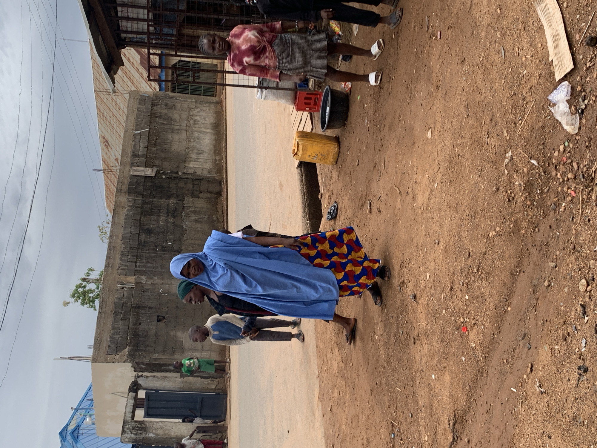The image depicts a street scene in Akwanga, Nigeria. The environment consists of a dirt ground and a paved road bordered by buildings. One building appears to be made of concrete while the other is composed of corrugated metal. The sky is overcast.
Several people are visible. An older woman stands near a road with a container and a red crate. A woman wearing a blue head covering and colorful dress walks toward the right of the image. A small group of people, including children, stand or walk near a building in the background.