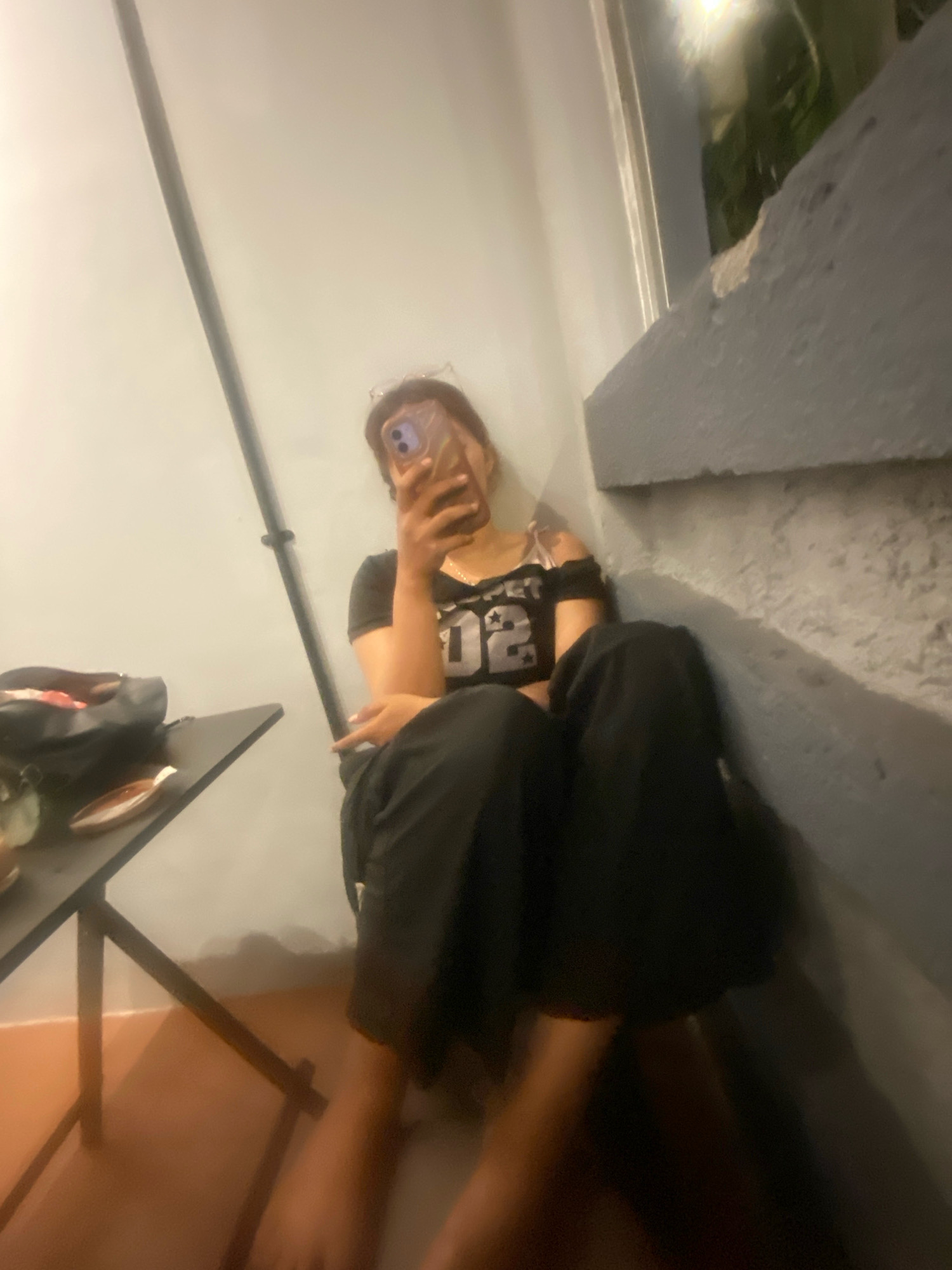 A person is taking a selfie with a phone in their hand, sitting in a corner of a room. The person is wearing a black off-the-shoulder top with "02" printed on it in silver, and black pants. A black bag and a small table are visible to the left of the person. The walls of the room are light-colored and grey. The floor is a warm brown color. The overall lighting is dim, with some light sources casting shadows. The image does not provide any specific information or context to determine the location as Klurak, Indonesia.