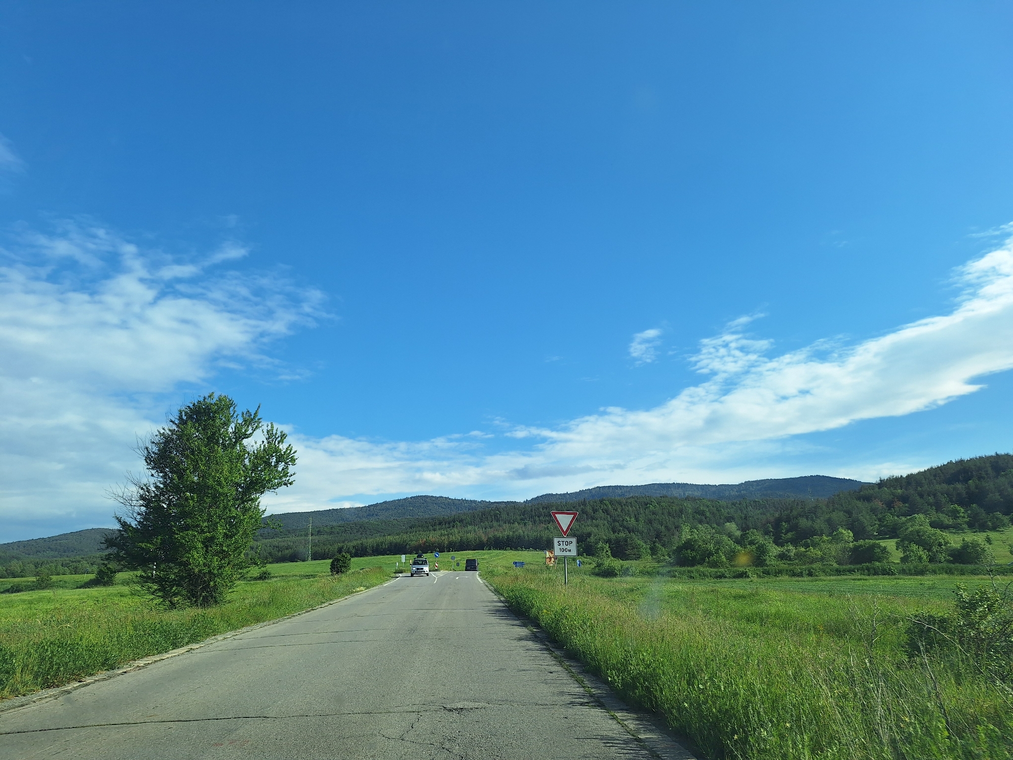 A daytime shot of a two-lane asphalt road cutting through a grassy landscape near Belchinski Bani, Bulgaria. A tree stands on the left side of the road, and a triangular "Yield" sign is visible on the right. Two vehicles are present on the road in the distance. Hills covered in trees are visible in the background under a blue sky with scattered white clouds.