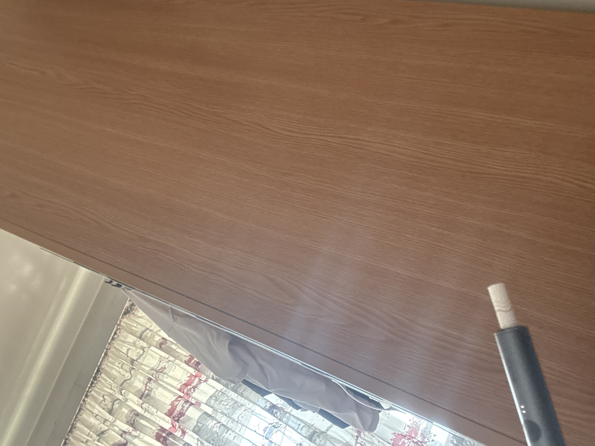 A low-angle, close-up shot shows a light-brown, wood-grain surface dominating the frame. In the lower right corner, a gray cylindrical object with a lighter-colored tip is visible. Below the wood-grain surface, at the left and bottom of the frame, a patterned shower curtain with white, red, and gray colors hangs. The location and details suggest an indoor setting, likely a bathroom. No people are visible. The location in the city of Troyan, Bulgaria is not evident from the image.