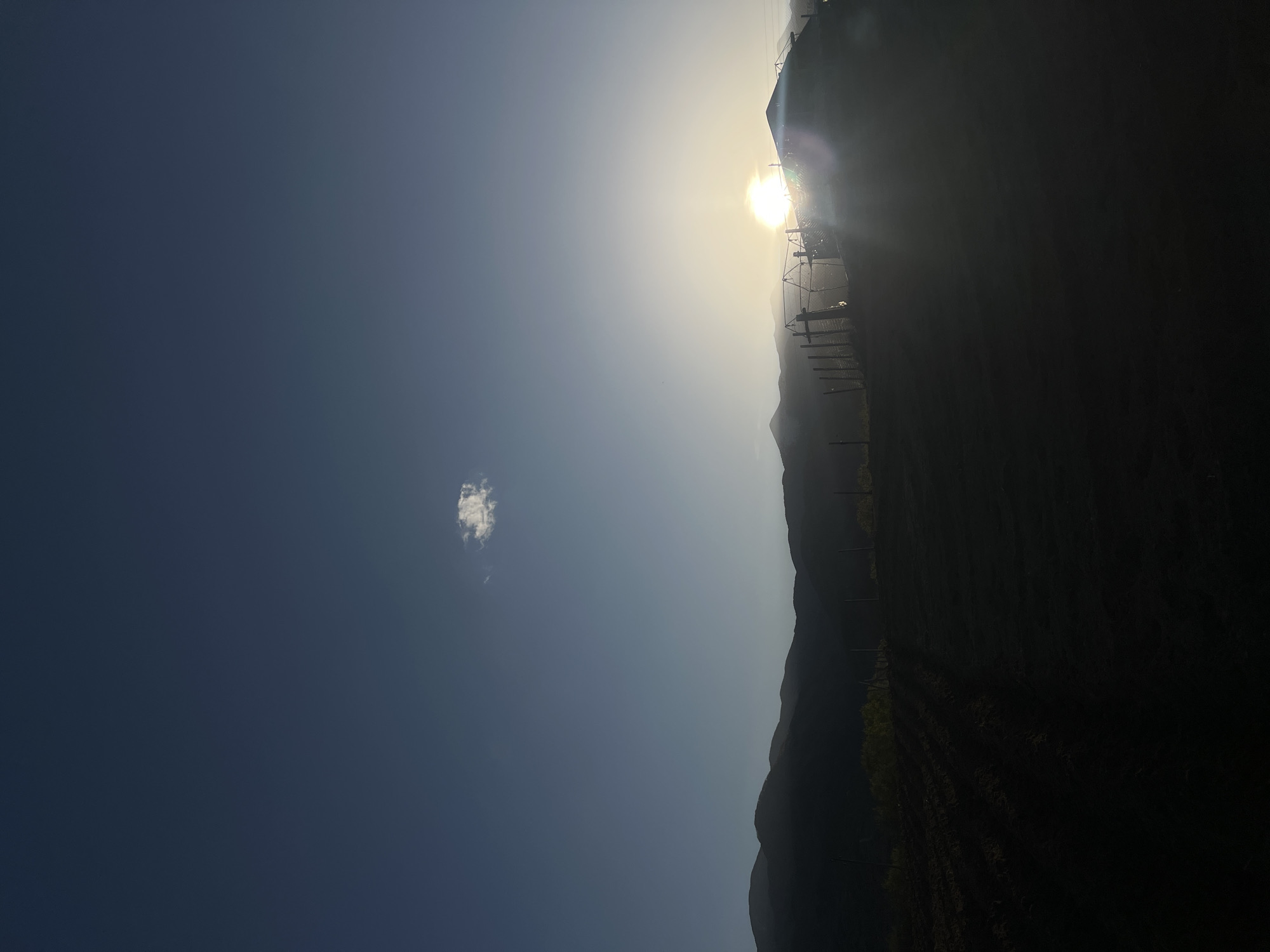 The image depicts a bright, sunny landscape, originally captured in a landscape orientation but presented here rotated 90 degrees clockwise. The dominant feature is a clear, deep blue sky filling the majority of the frame, with the sun positioned high, creating a powerful glare and lens flare in the upper right quadrant of the correctly oriented image. This strong backlighting silhouettes the foreground and midground elements.
A single, small, wispy white cloud is visible in the left portion of the blue sky. Below the horizon line, a dark silhouette of rolling mountains or hills stretches across the scene. In front of these mountains, closer to the viewer, are indistinct structures, possibly power lines, agricultural trellises, or scaffolding, also silhouetted against the bright sky. Further down, rows of dark vegetation or crops can be faintly discerned in the shadowed foreground, suggesting cultivated land.
The time of day appears to be mid-day or early afternoon due to the sun's high position and intense brightness. The weather is clear and sunny, though the strong sunlight creates a hazy atmosphere around the sun itself. No people or specific activities are visible. There is no discernible text in the image. The scene conveys a sense of a bright, vast outdoor space in a mountainous or hilly region, consistent with the provided location context of Kornitsa, Bulgaria.