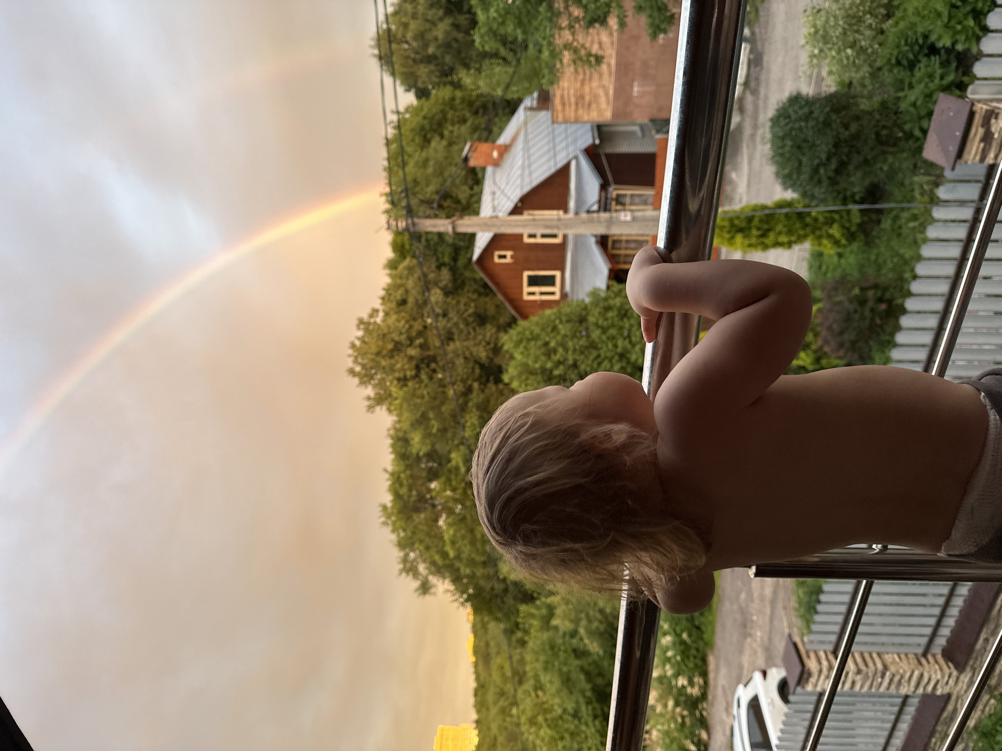 A child stands on a balcony, facing away from the camera, looking at a double rainbow in the sky. The child's back is to the viewer, and their hair is light brown and somewhat disheveled. They are wearing what appears to be a diaper or shorts. The balcony has a metal railing. In the background, there are lush green trees, a red-roofed house with several windows, a utility pole with wires, and a distant tall building. The sky is a warm orange and yellow hue, indicating either sunrise or sunset, with clouds present. The overall scene suggests an outdoor environment, possibly a residential area within a city. Without additional context or identifying markers, it is not possible to definitively confirm the location as Nizhny Novgorod, Russia.