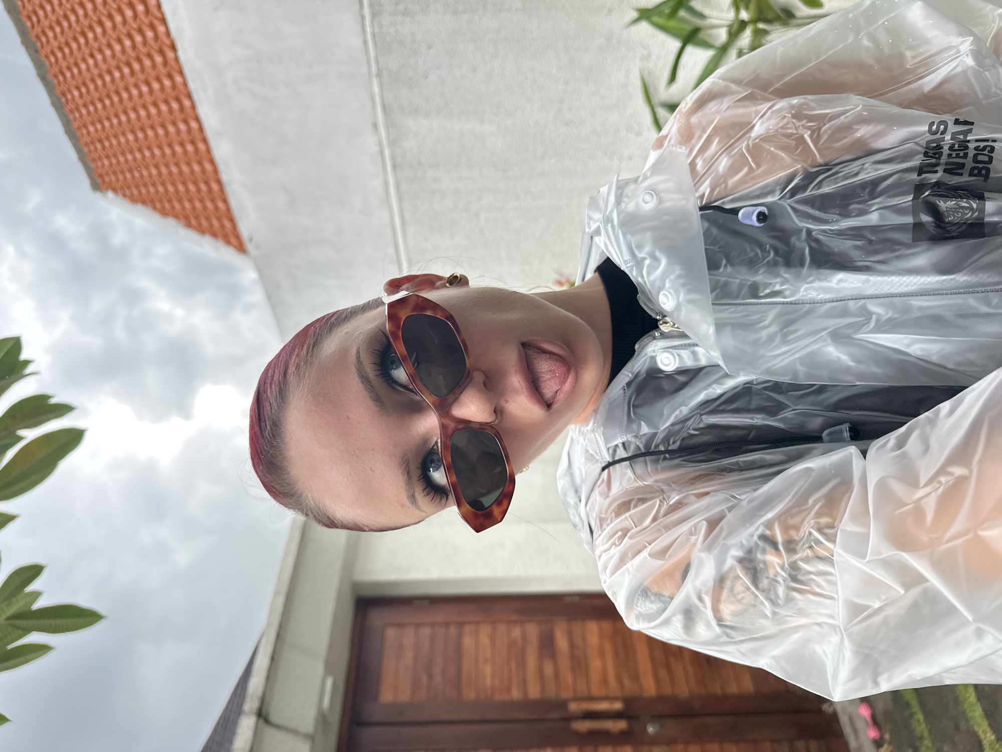 A close-up, rotated portrait captures a young individual with reddish-brown hair, playfully sticking their tongue out. They are wearing large tortoiseshell-patterned sunglasses and a clear plastic raincoat, fastened with white snaps, over a black top. Visible through the translucent raincoat are white text that reads "TUAS NEGAT BOS!" and a dark emblem or logo on the black top. The person looks directly at the camera with a mischievous and lighthearted expression.
The background is dominated by an overcast, cloudy sky. To the viewer's left, green foliage peeks into the frame. To the viewer's right and below the subject, a building is visible, featuring an orange, textured roofline, a white wall, and a prominent wooden door. The weather appears dull and overcast, suggesting it might be rainy, fitting the use of the raincoat. The image is rotated 90 degrees clockwise, giving it an unusual, tilted perspective. The setting is outdoors in North Kuta, Indonesia, likely during daytime.