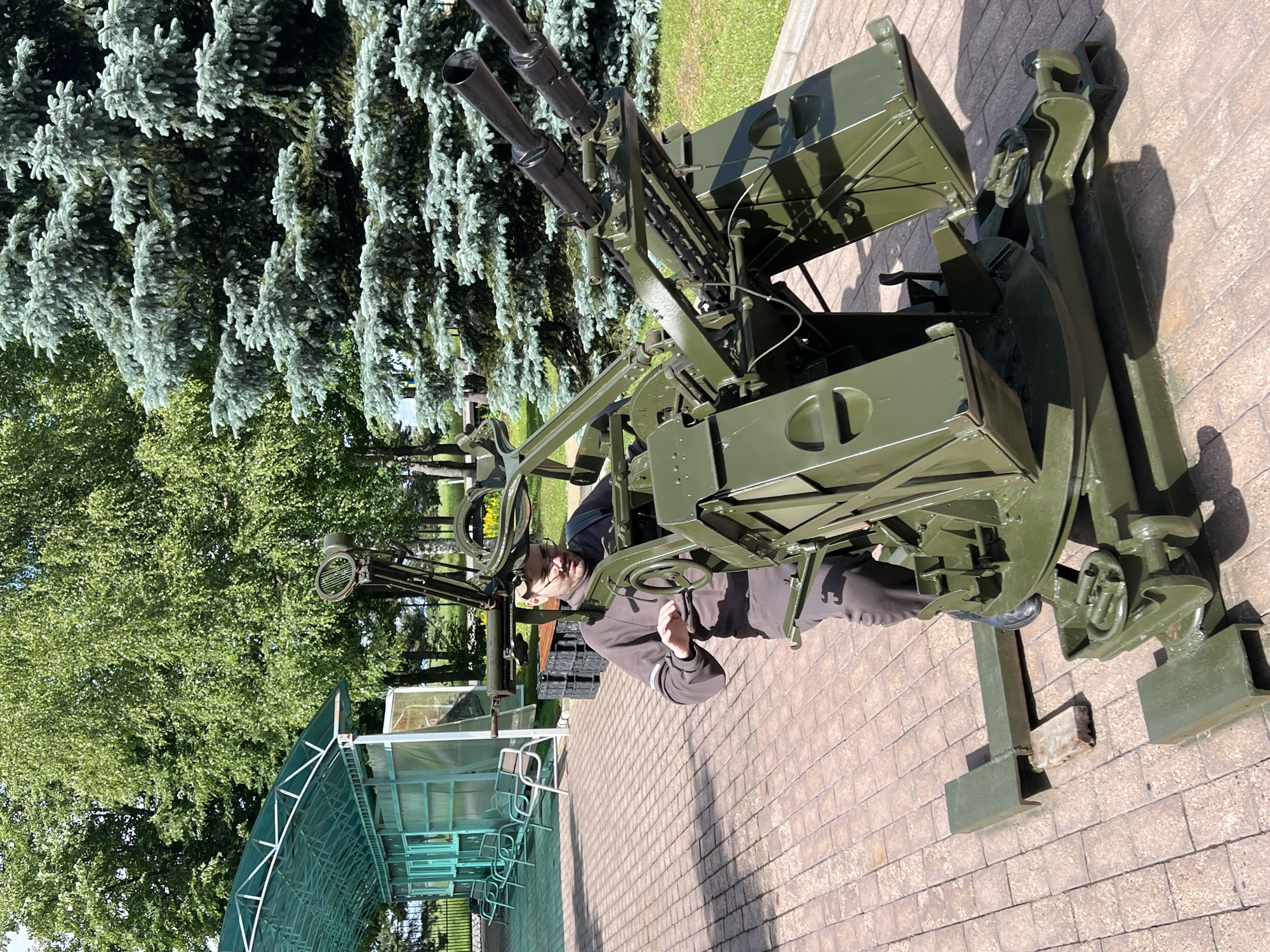 The image shows a man in a grey hoodie and glasses interacting with a military anti-aircraft gun. The gun is dark green and appears to be mounted on a tripod. It has multiple barrels and a complex mechanical structure. The man is positioned as if he is operating or inspecting the gun, with his hand on a steering wheel-like mechanism. He is looking towards the gun with a neutral expression.
The setting is outdoors, with a paved brick walkway in the foreground and middle ground. Behind the gun and to the left, there are large evergreen trees, possibly spruce, with characteristic needle-like foliage. Further back, there are more deciduous trees with full green canopies, indicating a daytime scene in a temperate climate, likely during summer. In the background, there's a glimpse of a green lawn and a covered structure with a metal frame and a translucent roof. The sunlight suggests it is a clear day.
The overall scene appears to be an outdoor exhibition or museum, possibly at a military history site, given the presence of the anti-aircraft gun. The lighting and shadows suggest it is midday. There is no visible text in the image. The focus is on the man and the detailed mechanism of the gun.