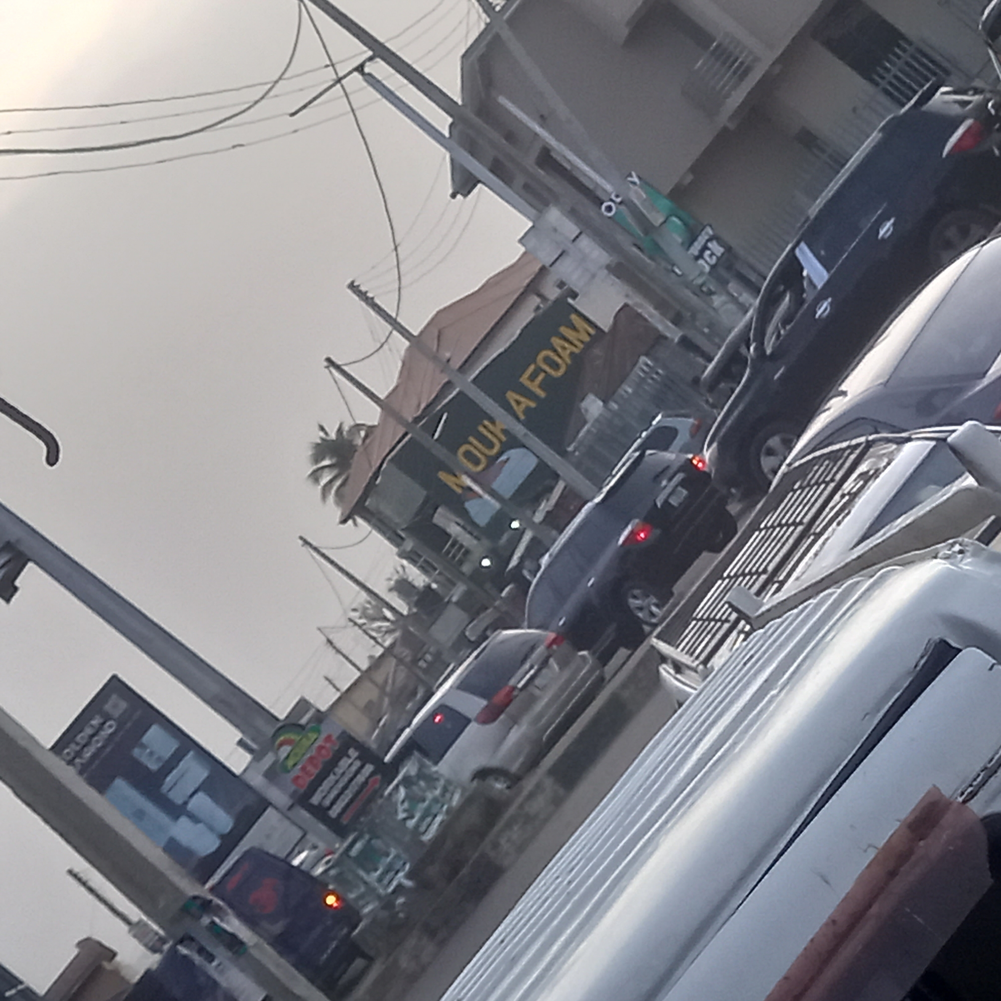 The image presents a tilted view of a street scene, likely in Dadi Riba, Nigeria, during what appears to be an overcast or hazy day. The perspective suggests the photo was taken from inside a vehicle, with the roof of a white car visible in the immediate foreground, indicating the photographer is in traffic.
The scene depicts a multi-lane road with several vehicles. A dark blue or black sedan is prominently featured in the middle ground, followed by a silver hatchback and another dark SUV further back. All cars appear to be moving slowly or stopped in traffic.
Along the roadside, commercial buildings and signs are visible. On the left, a large black billboard displays the partially visible text "GOLDEN DIAMOND" at the top. Below it, another sign shows the word "DEPOT" and some smaller, less legible text that includes "WORKABLE." On the right, a significant green building features a large sign with yellow lettering that clearly reads "MOUKA FOAM," suggesting a business selling foam products. Utility poles with multiple overhead wires crisscross the sky, and palm trees can be seen behind the buildings, indicating a tropical environment. The sky is a uniform light grey, consistent with an overcast condition. No individuals are clearly visible in the frame.