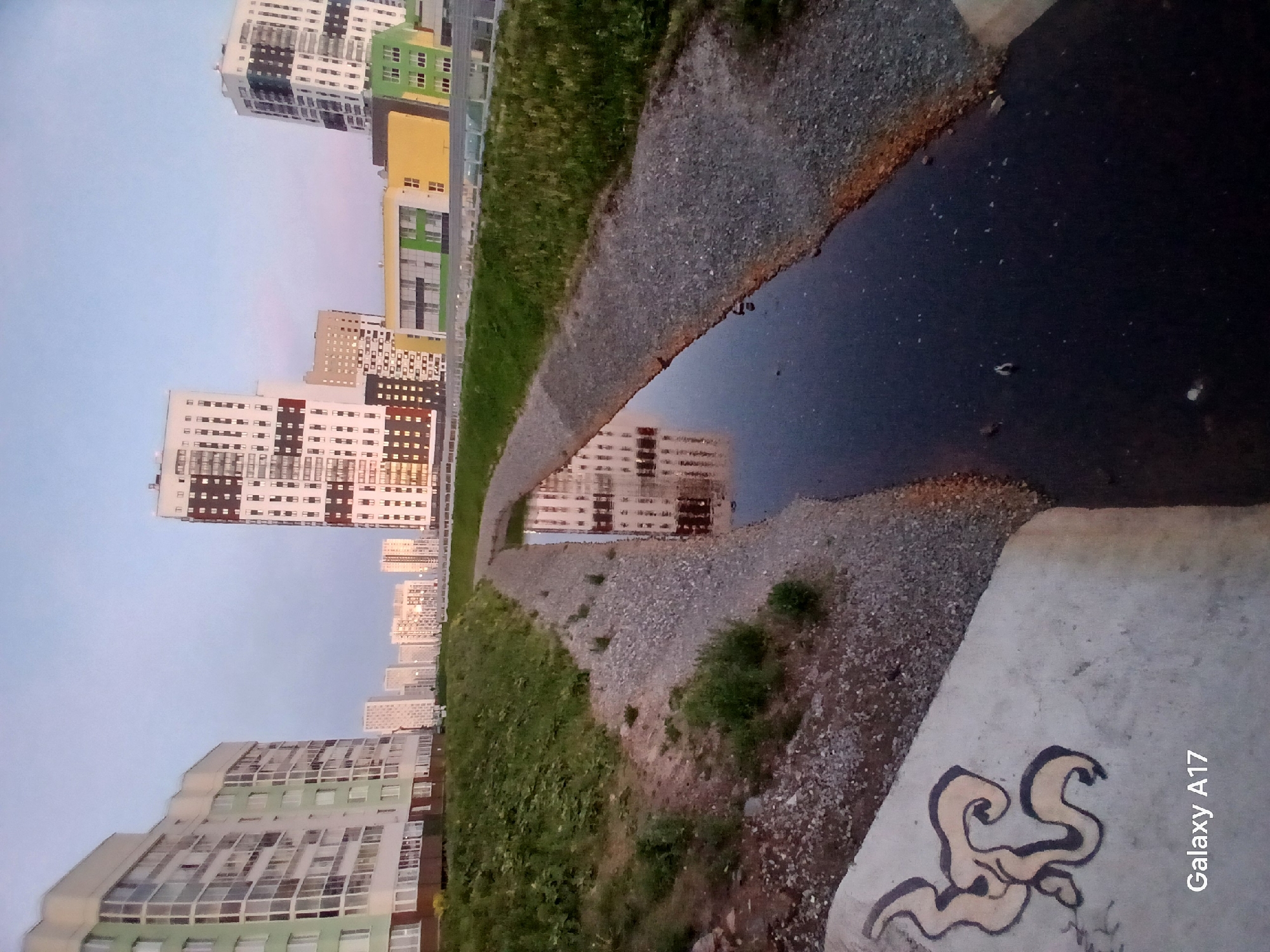Here's a description of the image:
**Scene Overview:** The image shows a water-filled drainage ditch alongside a sidewalk in an urban area. The viewpoint is low, looking along the ditch.
**Environment:**
* **Location:** Yekaterinburg, Russia (based on the buildings and overall appearance).
* **Urban area:** The scene includes tall residential buildings in the background, indicating a densely populated area.
* **Drainage ditch:** A narrow, dark, water-filled ditch runs along the sidewalk.
* **Sidewalk:** The sidewalk is concrete, with graffiti on one edge.
**Objects:**
* **Buildings:** High-rise buildings with different colors and architectural styles line the background.
* **Gravel:** Small gravel is scattered on the edges of the ditch.
* **Grass:** A strip of grass borders the gravel and ditch.
* **Water:** The ditch is filled with dark water, reflecting the buildings.
* **Graffiti:** A light-colored design is painted on the edge of the concrete sidewalk.
**Actions/Interactions:**
* **Reflection:** The buildings are reflected in the water, indicating calm conditions.
**Miscellaneous:**
* The photo was apparently taken by a "Galaxy A17" smartphone.