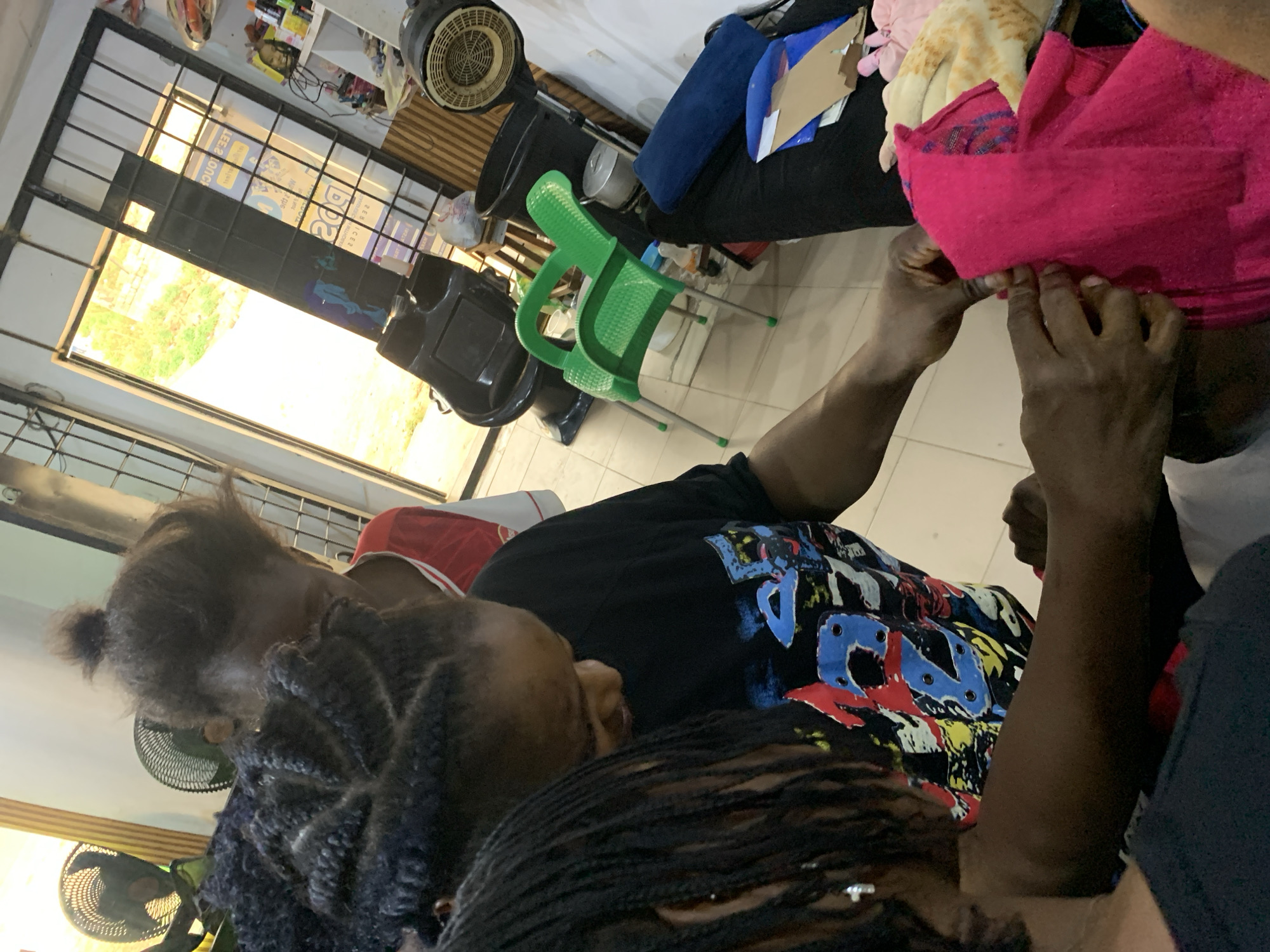 The image shows the interior of a salon in Jos, Nigeria. Three individuals are present. One person, partially visible, has their head wrapped in a pink towel and is attended to by two other people who are working on their hair. The environment appears busy, with a hair dryer, salon chair, and various products and supplies visible in the background. A window provides a view of the outside, suggesting it's daytime.