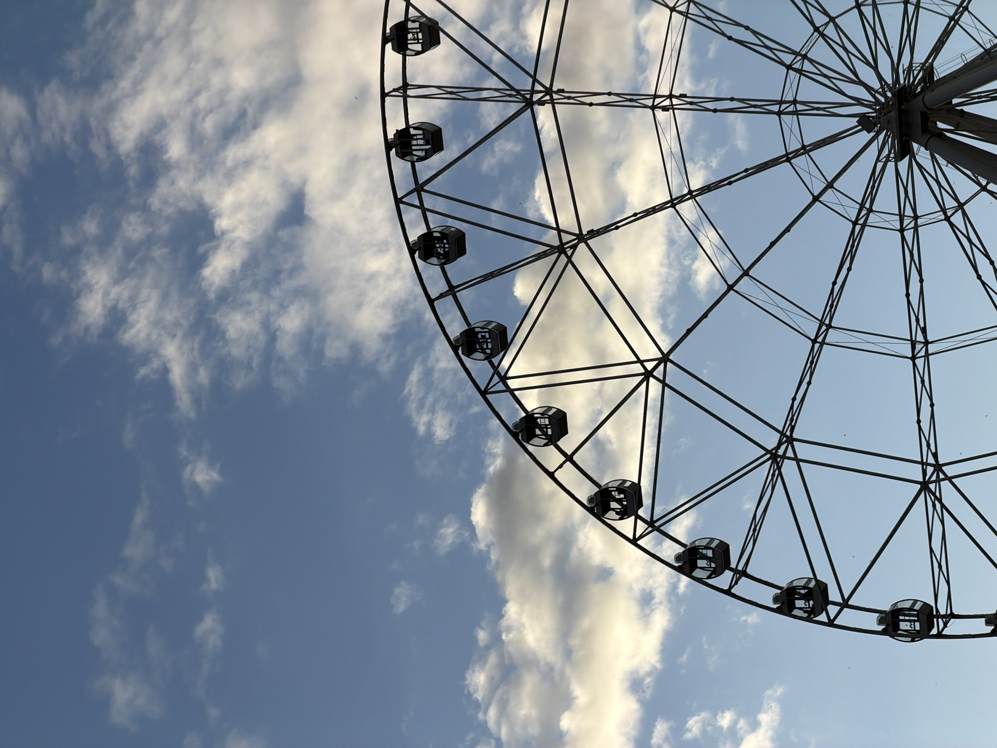 The image shows a partial view of a Ferris wheel against a partly cloudy blue sky. The wheel's frame is visible, along with several passenger cars. The image is taken from a low angle, looking up at the wheel and sky. The location is Rostov-on-Don, Russia, based on the presence of a Ferris wheel, which is a tourist attraction there.