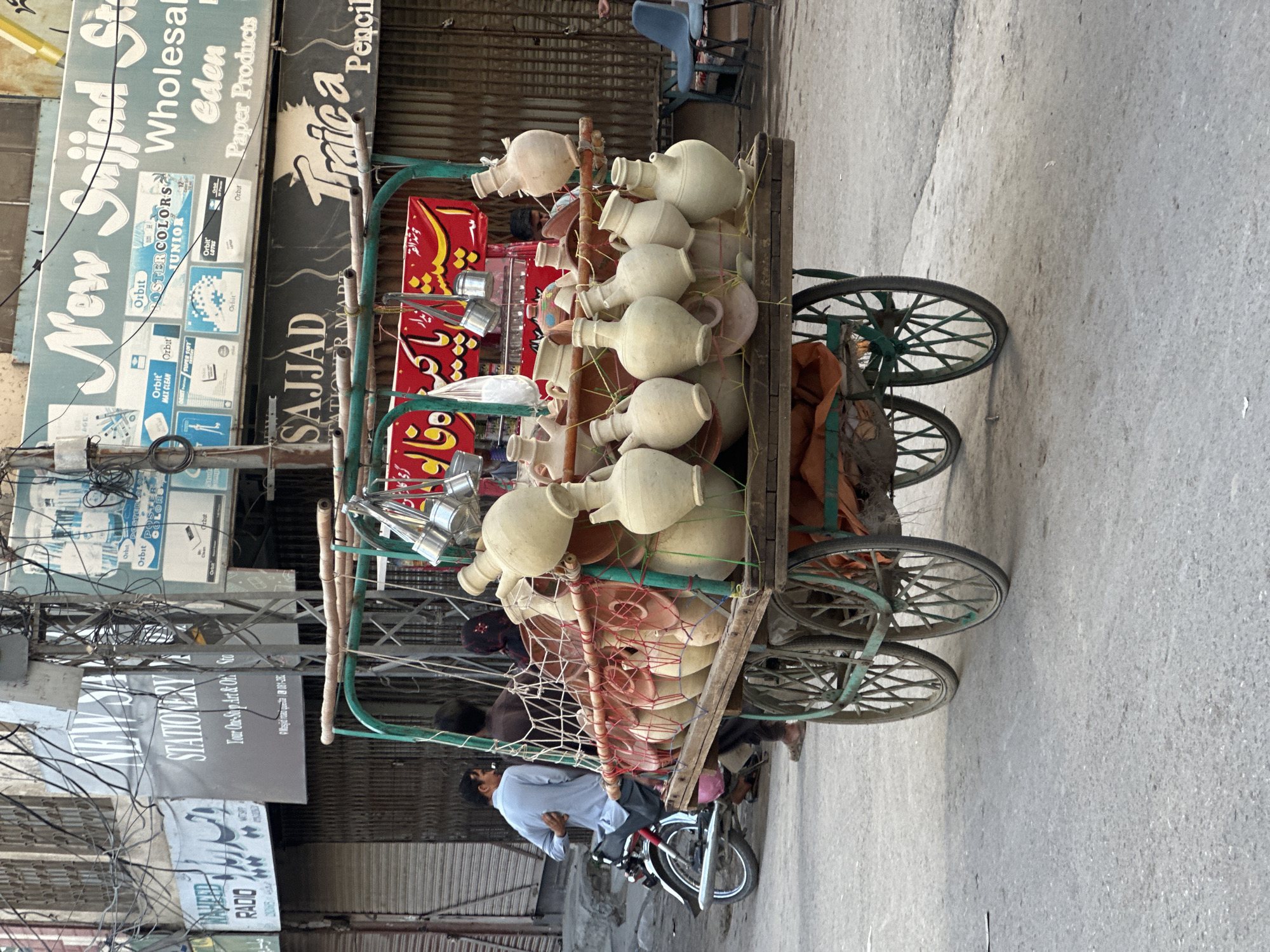 The image portrays a street scene in Quetta City Tehsil, Pakistan, featuring a traditional cart laden with clay pots. The cart, possibly for selling water or beverages, is parked near commercial establishments with signs in Urdu and English. A man is riding a motorcycle nearby. Overhead are a web of electrical wires, typical of urban areas. The overall impression is of a busy, perhaps somewhat congested urban environment.