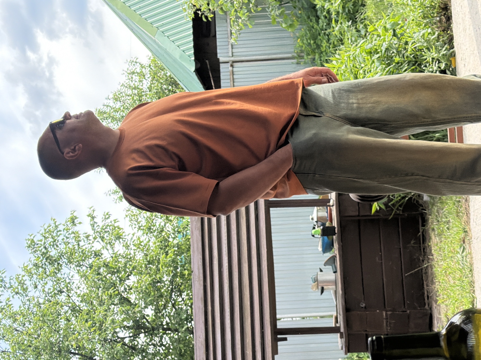 A man stands outdoors, looking upwards towards a bright, cloudy sky. He is bald, wears sunglasses, a rust-orange short-sleeved t-shirt, and olive green trousers, with one hand casually in his pocket. To his left, green trees and bushes are visible, with parts of a building featuring a green corrugated metal roof and a wooden slatted structure. To his right, a dark wooden table or counter holds various metallic kitchen pots and utensils. The ground is a mix of green grass and a paved surface. A dark glass bottle is partially visible in the bottom right foreground. The scene depicts a relaxed daytime moment in what appears to be a garden or backyard setting, likely during a warm season, potentially in Chaikovsky, Russia. No text is visible.