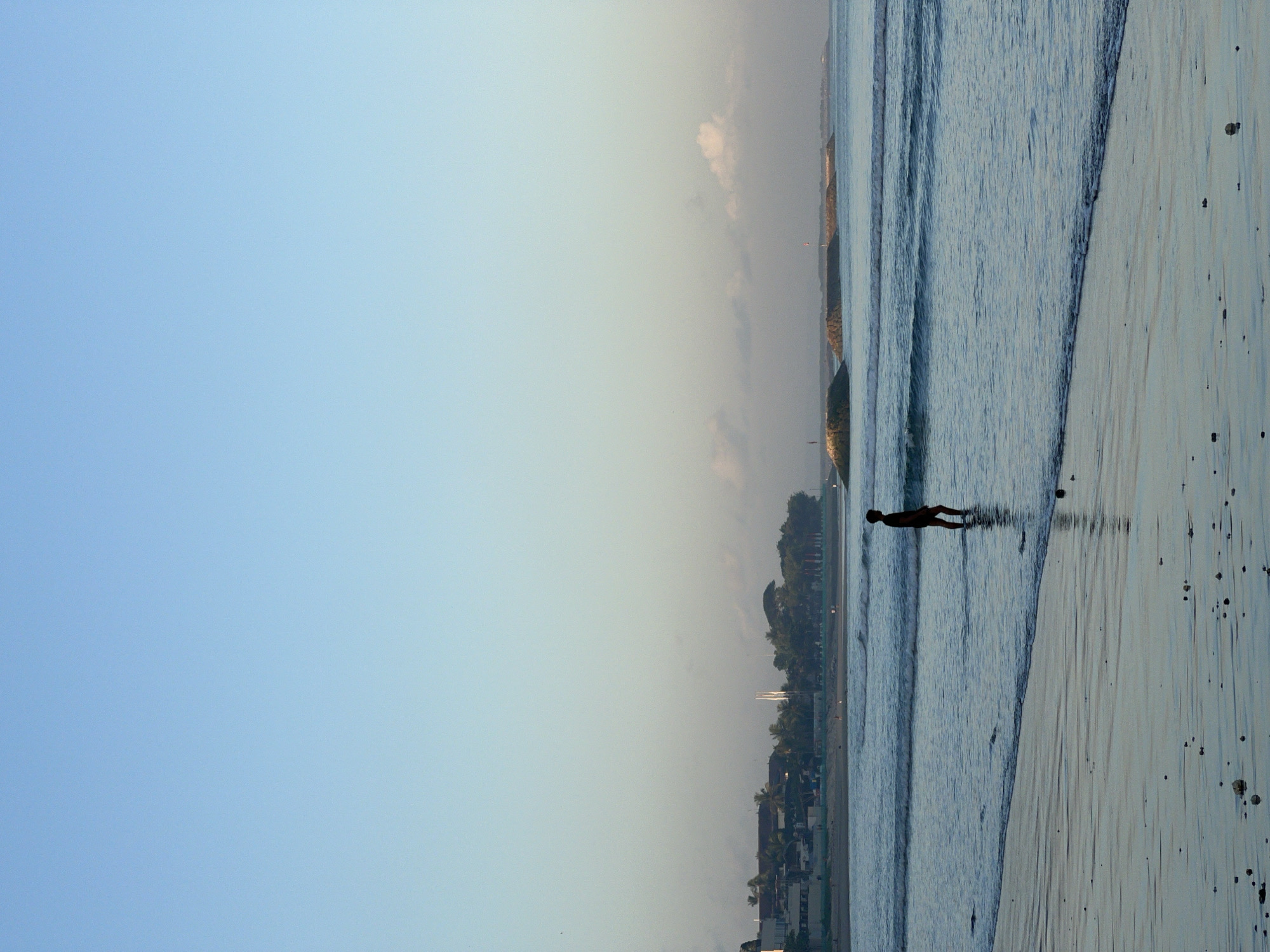 A lone figure stands in the shallow ocean water at Kuta Beach, Bali, Indonesia. The figure is silhouetted against the gentle waves and the sandy shore. In the background, the coastline is visible with buildings and palm trees. The sky is a clear, pale blue, with a few wispy clouds near the horizon. The water reflects the soft light of what appears to be early morning or late afternoon. The beach itself is covered with small stones and debris.