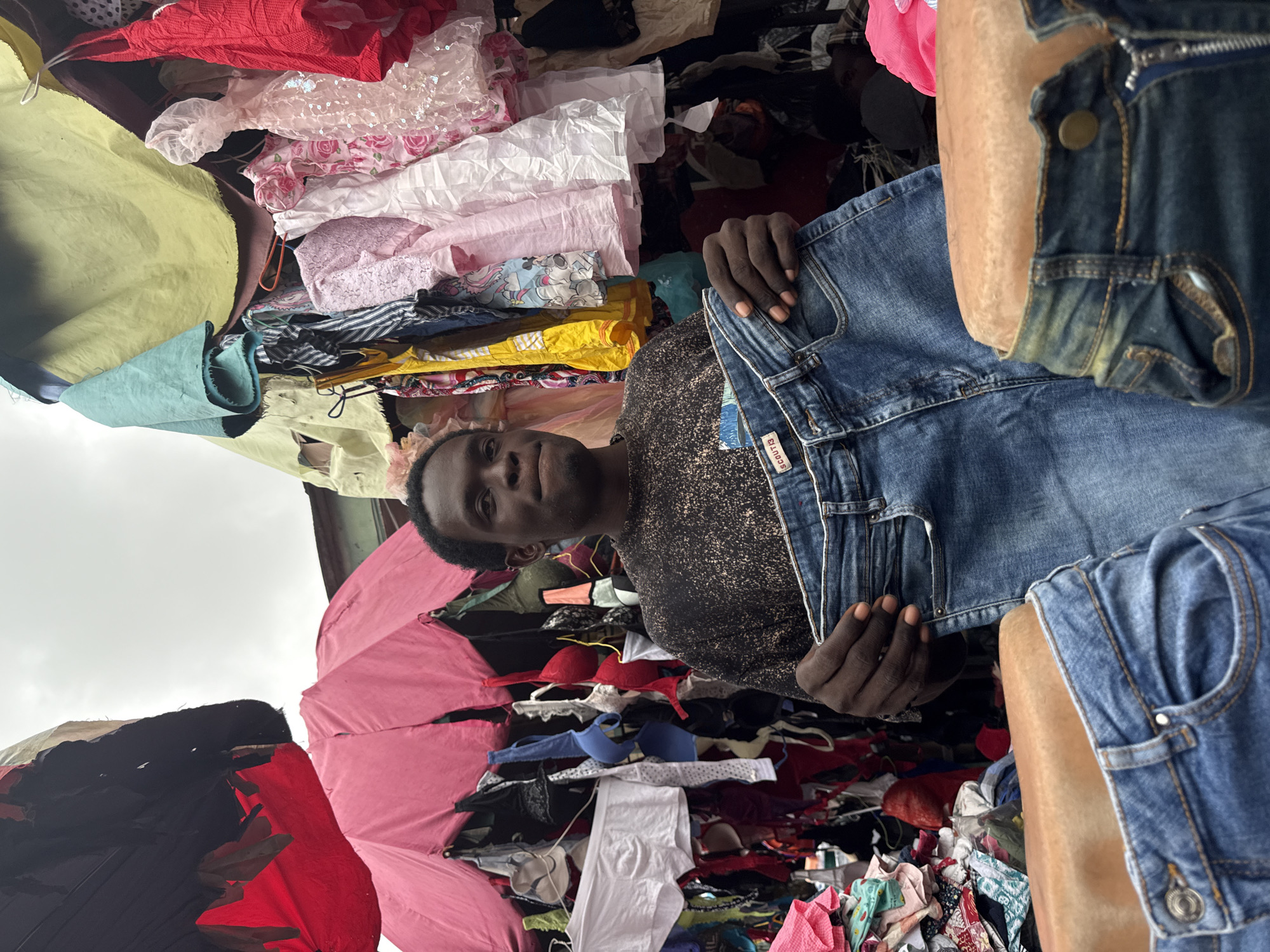 In an open-air market stall in Lagos, Nigeria, a young Black man stands holding up a pair of blue denim jeans. He is wearing a dark, speckled long-sleeved shirt. The stall behind him is packed with a colorful array of clothing, including bras, shirts, and other garments, suspended from various fixtures under awnings made of fabric. In the foreground, the waistbands and upper portions of two pairs of jeans are displayed on mannequins. The overall scene suggests a busy retail environment for apparel.
