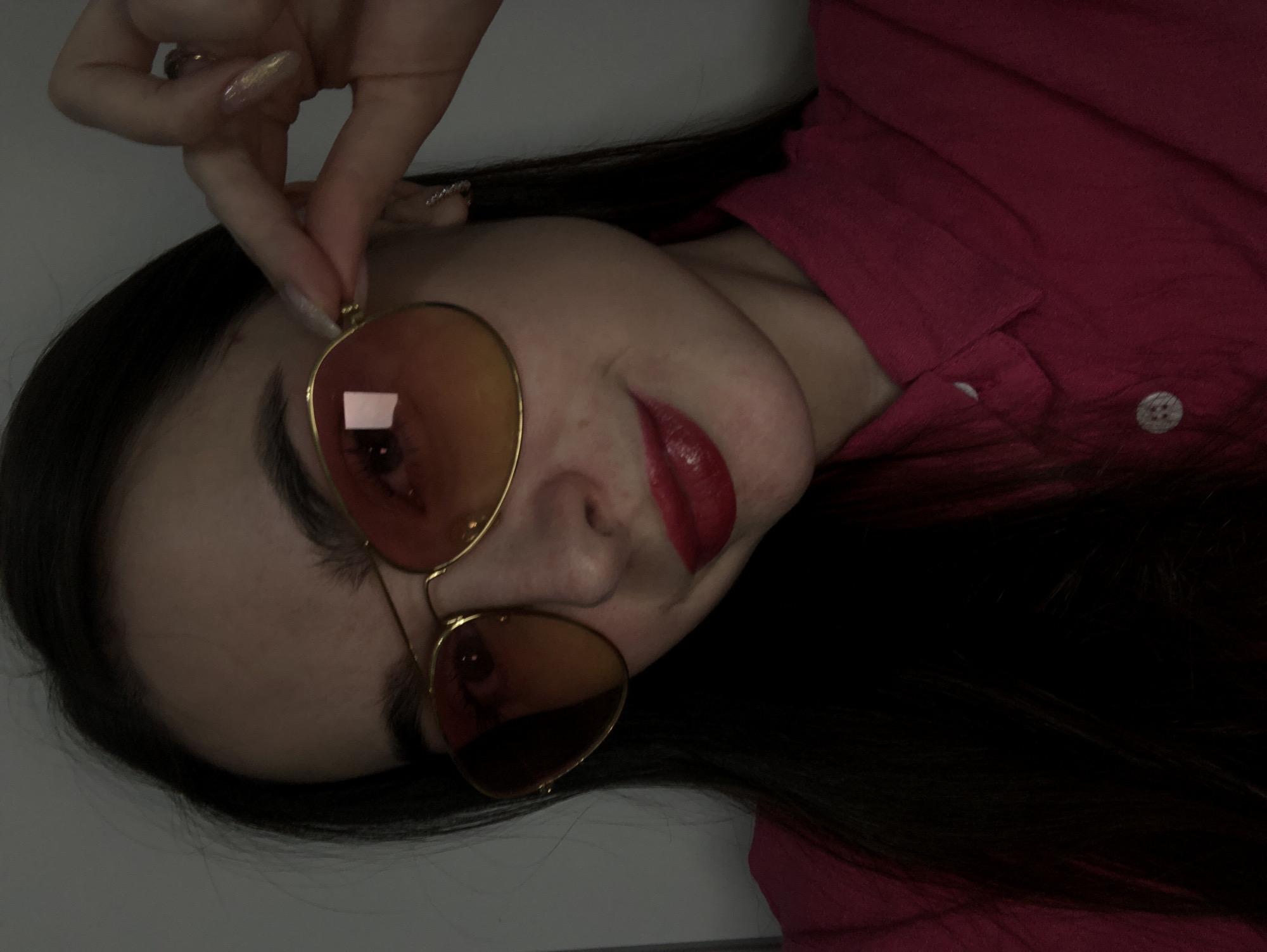 A woman with long dark hair and fair skin is posing for a selfie. She is wearing a pink shirt, red lipstick, and gold-rimmed sunglasses that she is holding with her left hand. The background is a blurry white. No information suggests the image was taken in Noviy, Russia.