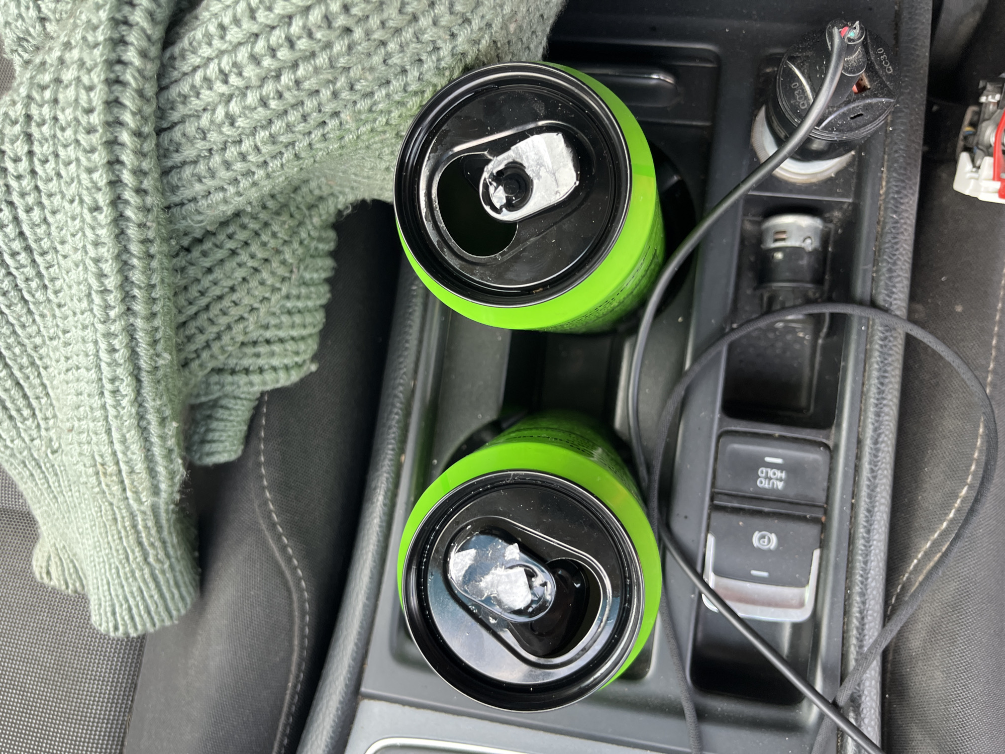 The image shows two open cans of a bright green beverage sitting in car cup holders. A knitted green sweater is draped over the car's interior. In the foreground, there are car controls, including an electronic parking brake button and an "AUTO HOLD" button, as well as a car charger plugged into a power outlet. The scene appears to be a stationary vehicle, possibly parked. There are no people visible in the image. The location is identified as Vasilyevo, Russia.