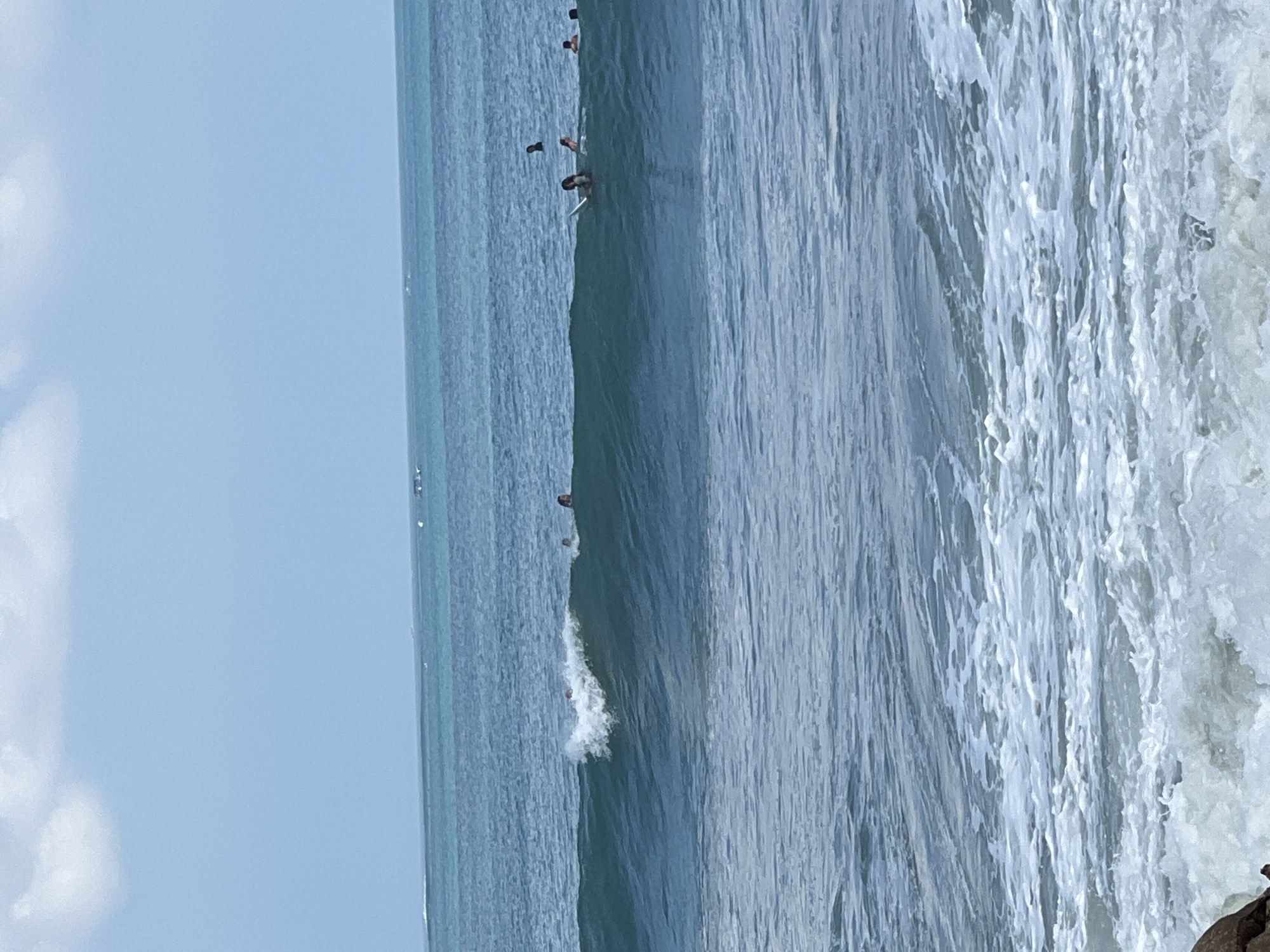 In the ocean off the coast of Kuta, Indonesia, surfers are riding waves under a bright, partially cloudy sky. The water is a deep blue, with lighter turquoise hues closer to the horizon. Small whitecaps are visible on the surface. In the foreground, foamy waves crash onto the shore, with a large dark rock visible on the left. Several surfers are scattered throughout the water, some riding waves and others waiting for their turn. One surfer is on a surfboard, paddling through the water.