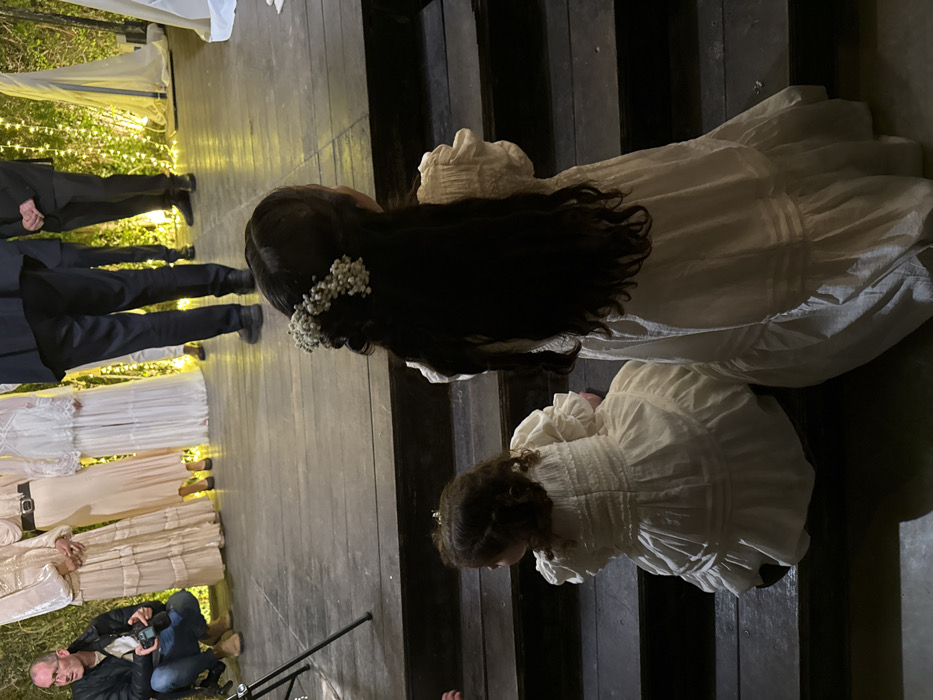 The image captures an outdoor evening scene, likely a formal event such as a wedding or a celebratory gathering, viewed from an elevated perspective. The main focus is on two individuals, an adult or older child and a younger child, both seen from behind and wearing white or cream-colored long dresses.
The person in the foreground, positioned higher up on what appears to be a wooden platform or steps, has long, dark, wavy hair adorned with a cluster of small white flowers, resembling baby's breath, on the left side of their head. Their dress is light-colored, featuring a textured or pleated bodice and flowing fabric. Below and slightly to the right, a younger child is partially visible, seemingly kneeling or sitting on a lower step. This child also has curly brown hair and is wearing a similar white or cream dress with a textured bodice and puffed sleeves. Both individuals appear to be facing away from the camera, looking towards the left side of the frame.
The setting includes a wooden plank floor or decking that extends across much of the image. To the far left, several pairs of legs belonging to people in dark trousers and shoes are visible, suggesting other attendees, possibly men in formal attire. Further back on the left, other individuals wearing long, light-colored dresses can be seen. Bright string lights are draped in the background on the left, illuminating the area and indicating that the event is taking place at night or dusk. In the lower left corner, a man is crouched down, holding a camera, suggesting he is a photographer capturing the event. No discernible text is visible in the image. The overall atmosphere appears festive and intimate.