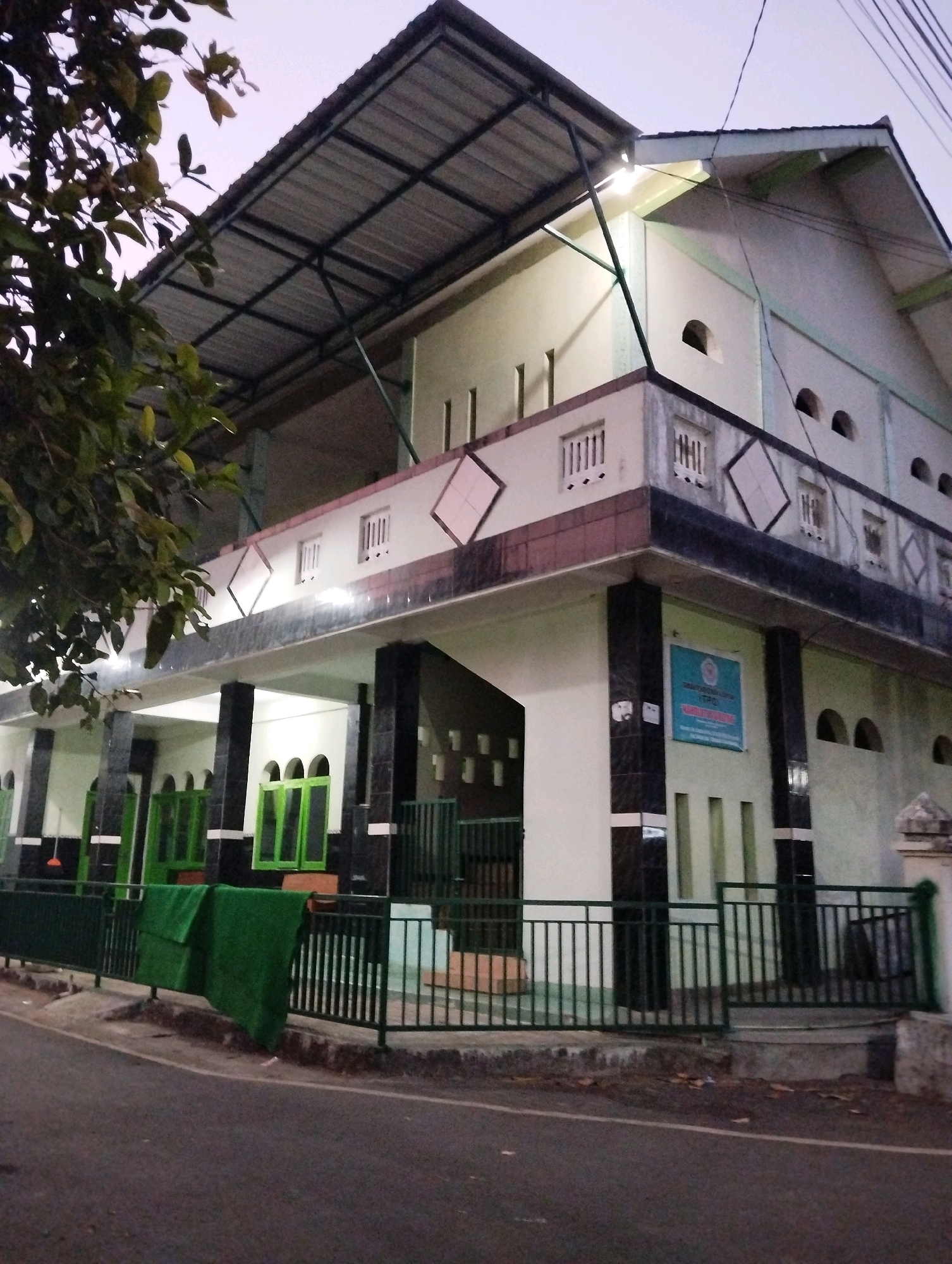 This is a low-angle, medium shot of a two-story building with a large, sloped metal roof supported by a metal frame. The building appears to be a public facility, possibly a mosque or a school, given the architectural style and the presence of a sign on the upper story. The exterior walls are painted a light color with dark trim and green accents. The lower level features several green-framed windows and doorways, some with arched tops. A dark metal fence runs along the front of the property, and a green tarp is draped over a section of it. The upper level has a balcony with decorative diamond patterns and small, barred windows. The building is located in an urban or semi-urban environment, with a paved road in the foreground. Trees with dark green foliage are visible on the left side of the image. The sky is overcast and appears to be dusk. The overall impression is that of a functional, well-maintained building in Salatiga, Indonesia.