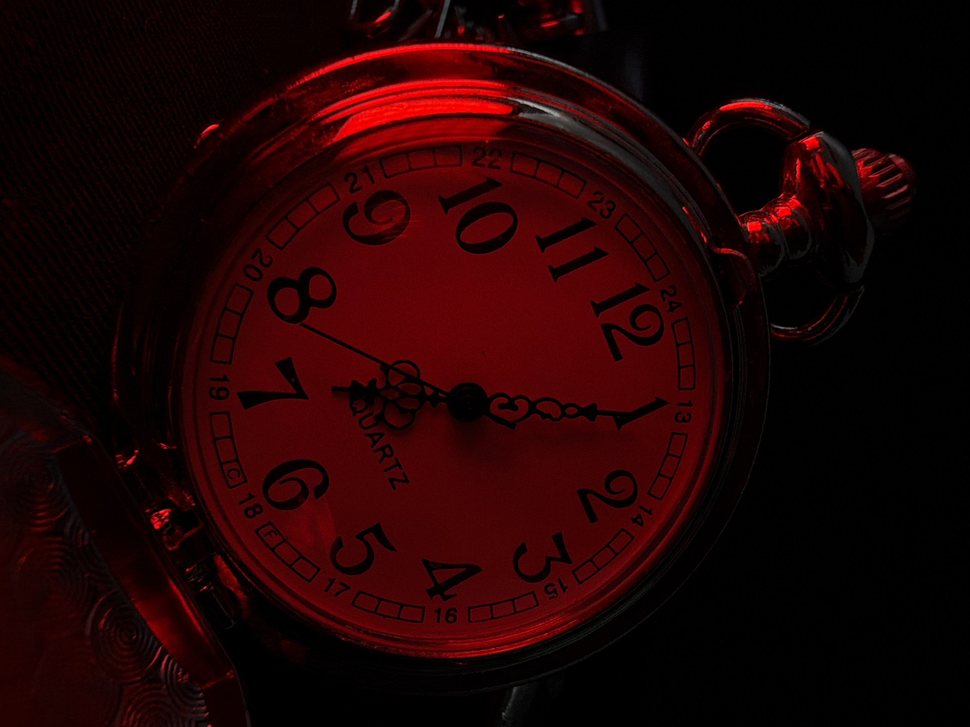 This is a close-up, low-angle, dark shot of a pocket watch illuminated by a red light. The watch face is round with black numbers and markings against a red background. The numbers visible include 2, 3, 4, 5, 6, 7, 8, 9, 10, 11, 12, 13, 14, 15, 16, 17, 18, 19, 20, 21, 22, 23, and 24. The hands of the watch are black and point to approximately 4:55. The word "QUARTZ" is visible on the watch face. The watch has a decorative outer case and a chain attachment mechanism that is also visible. The background is black, suggesting a dark environment or intentional lighting. The red illumination gives the image a dramatic and moody atmosphere. There is no discernible location context, time of day, or weather information. The image focuses solely on the detailed appearance of the pocket watch.