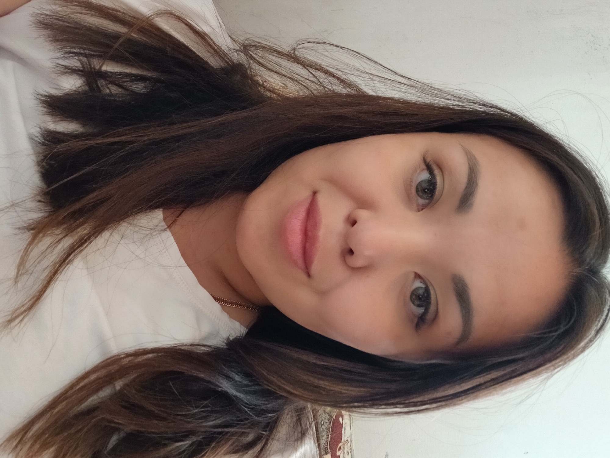 The image is a close-up, head-and-shoulders selfie of a young East Asian woman with long, dark brown hair. She is wearing a white t-shirt. Her eyes are a light brown, and she has a faint smile on her lips. The background is a plain, light-colored wall, with a hint of a patterned fabric visible on the right side of the frame. The lighting appears to be natural, coming from the front. The woman is positioned in the center of the frame, looking directly at the camera.