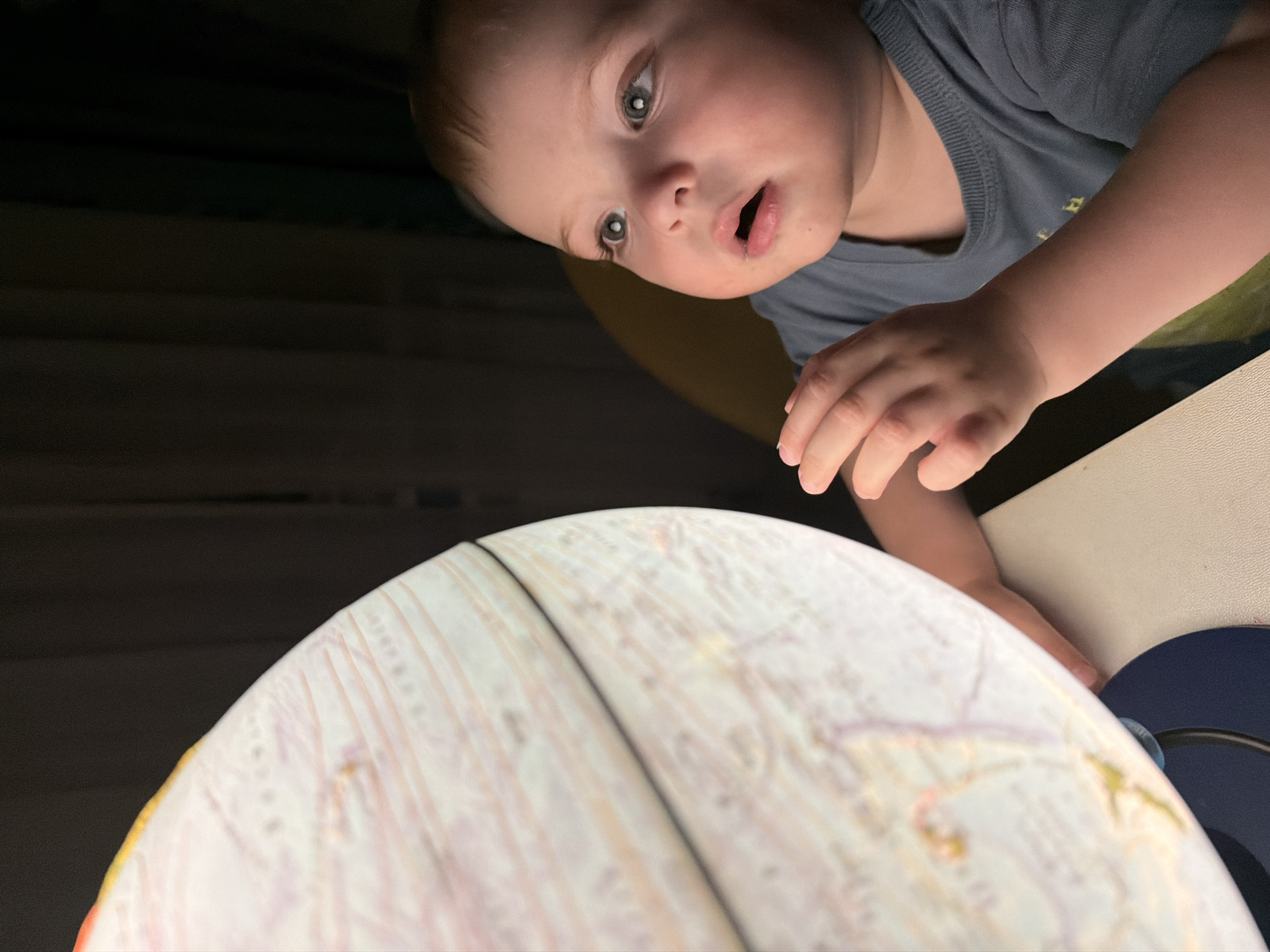 A young child with wide, curious eyes looks at a lit globe. The child is wearing a gray t-shirt. The globe is positioned on a table in front of the child, and its surface shows continents and countries. The background is dark and out of focus, suggesting an indoor setting. There is no visible indication of the specific location in Votkinsk, Russia.