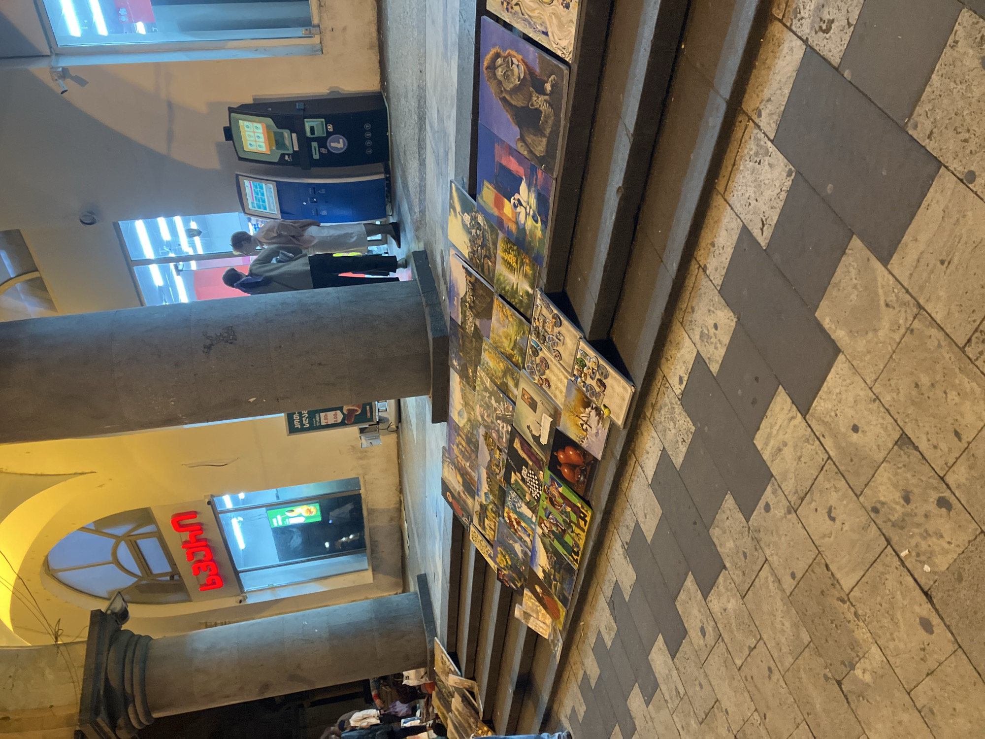 The media file depicts a street scene at night in Tbilisi, Georgia, illuminated by artificial lights. In the foreground on the right, numerous colorful paintings are displayed for sale on a set of wide, stone steps. The artwork features diverse subjects, including a prominent image of a lion, landscapes, and abstract designs. The ground is paved with a distinctive pattern of light and dark grey rectangular and square tiles.
On the left side of the image, two individuals are visible near two ATM-like machines. One machine has a screen displaying various icons, while the other features a circular "T" logo. Behind them, large arched windows with bright fluorescent lighting suggest storefronts. A substantial concrete pillar stands in the mid-ground, supporting an overhead structure. The building facade above the street features additional windows and architectural elements.
Visible text on signs confirms the location. A bright red neon sign reads "სამეფო" (Samepo), meaning "Royal." Another sign in a window mentions "ჯორჯია" (Georgia) and "ლარი" (Lari), the Georgian currency, alongside prices. Further left, another sign partially visible in a window reads "სინათლე" (Sinatle), meaning "Light." The scene suggests an active urban environment, possibly a market or commercial area, with people engaged in routine activities while a street vendor offers artwork.