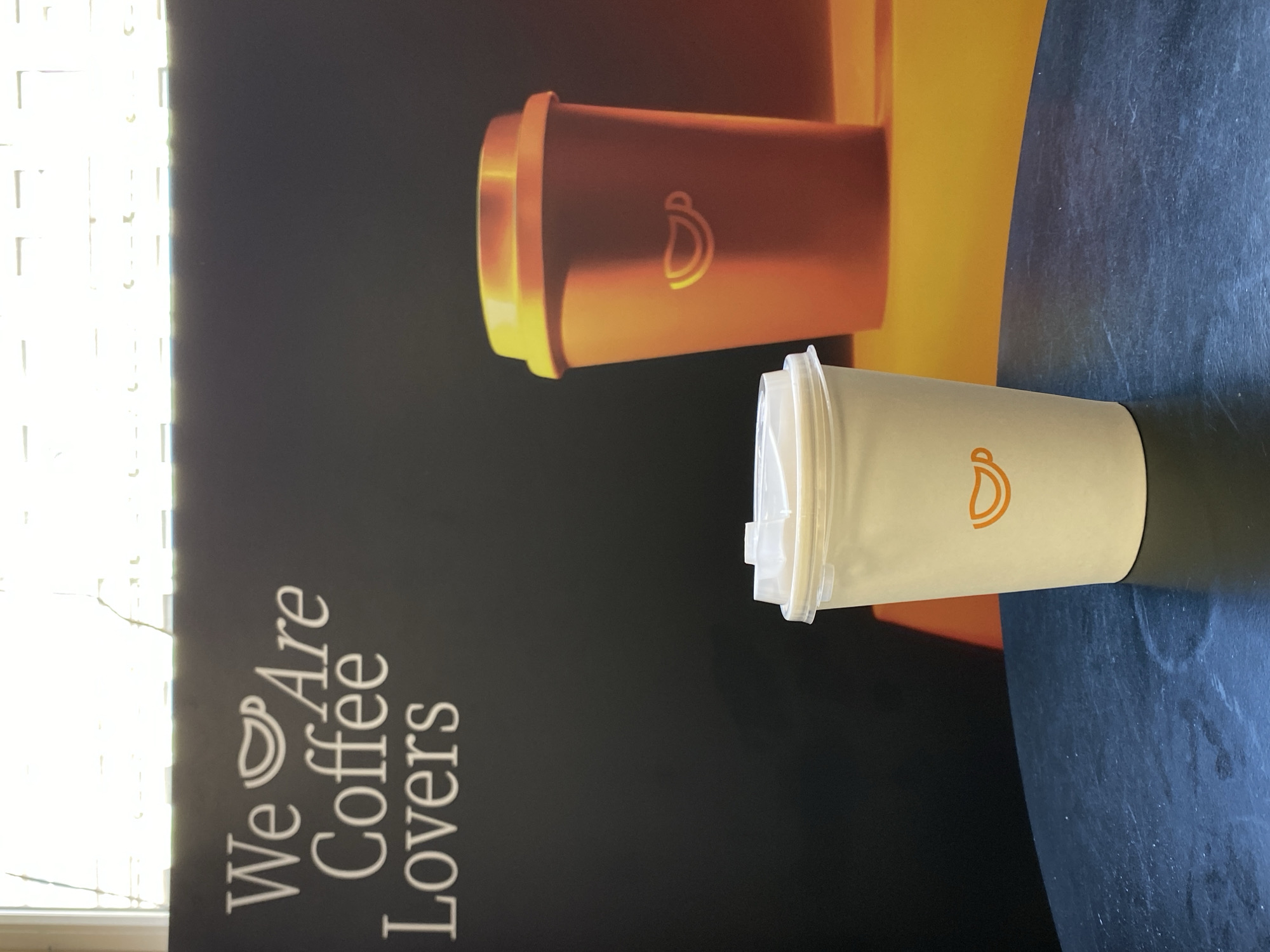 A coffee cup is lying on a black table in front of a wall advertisement that says "We Are Coffee Lovers". The cup is white with a plastic lid. The background consists of a colorful advertisement for coffee. The location in the city of Saint Petersburg, Russia is not evident from the picture.