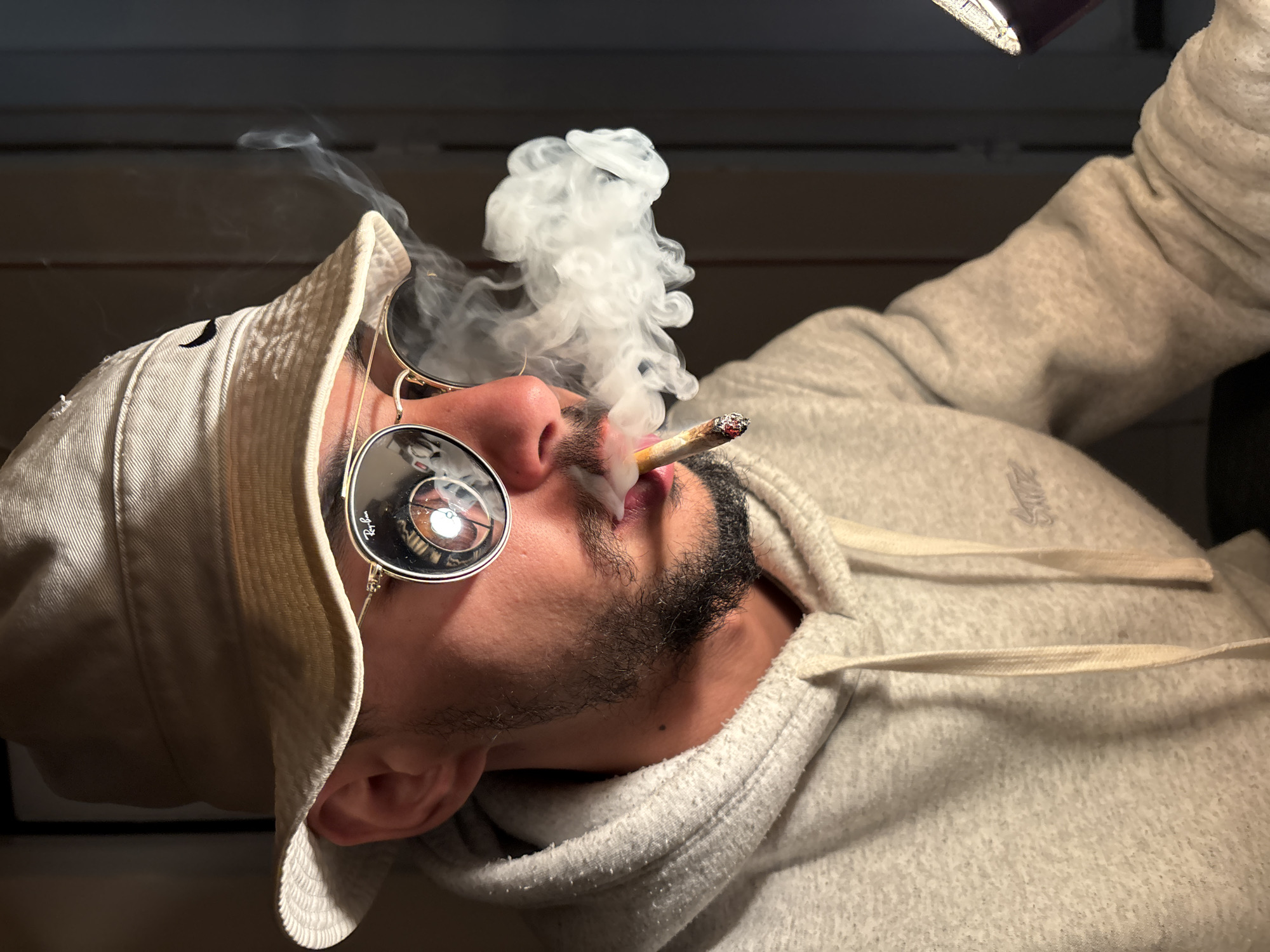 A single individual, a presumed male, is depicted in a chest-up, profile shot against a dark, featureless background, facing right. The person wears a light tan bucket hat with a black Nike swoosh logo, and gold-rimmed Ray-Ban aviator-style sunglasses. Reflections in the sunglasses show a bright circular light source and a purple object, possibly a phone case. The individual has a short, dark beard and is wearing a light grey speckled hooded sweatshirt with a faint logo on the chest. A hand-rolled cigarette, actively lit and glowing at the tip, is held between the lips, with a dense plume of white smoke exhaling from the mouth, drifting rightward. The lighting suggests an indoor environment. No specific location within the city of Tanger, Morocco, is discernible from the image.