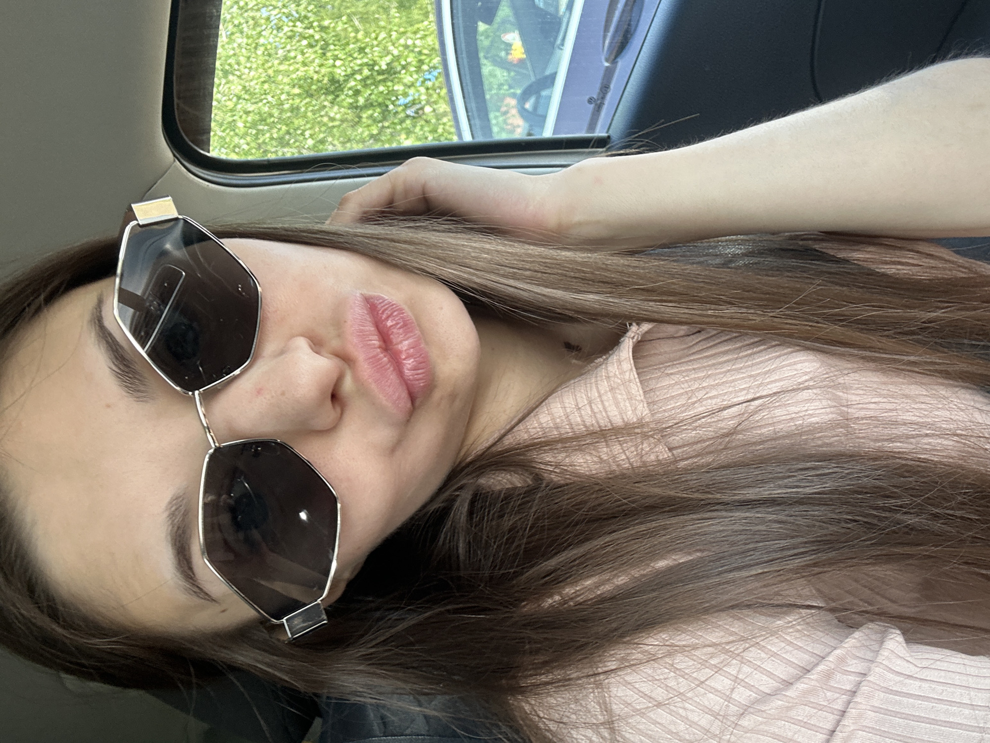 A selfie depicts a young woman inside a vehicle, presumably a car, as suggested by the window in the top left of the frame displaying greenery. The woman is wearing geometric sunglasses with a silver frame and dark lenses. She has long brown hair and is wearing a light pink, ribbed shirt. She is holding her hand near her forehead. The image's orientation is rotated. There is no information to suggest that the picture was taken in Хохряки, Russia.