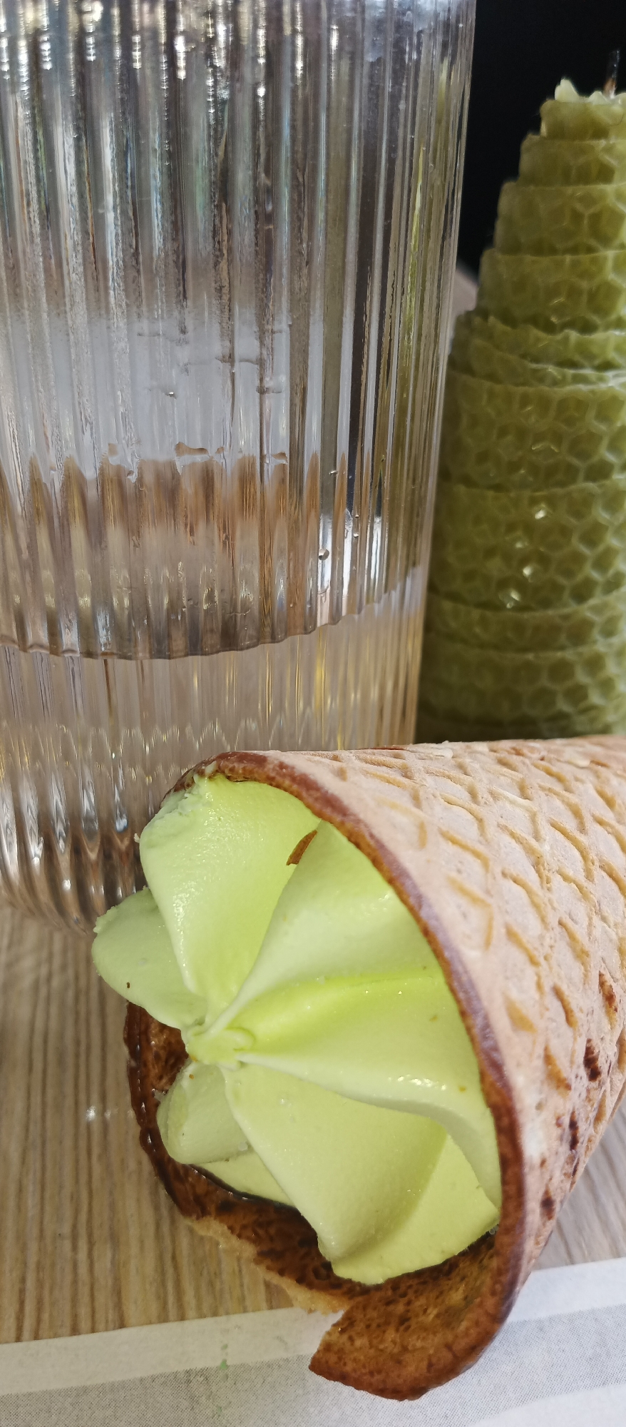 This close-up image captures a still life composition of refreshing treats and a decorative item, likely set indoors on a light-colored wooden surface.
In the upper left quadrant, a clear, fluted glass stands prominently. Its ridged surface exhibits condensation, suggesting it contains a cold liquid, possibly water. Light refracts through the glass, creating shimmering vertical lines and reflections.
Dominating the foreground, slightly off-center, is a waffle ice cream cone filled with light green soft-serve ice cream. The ice cream is swirled artfully, presenting a vibrant, smooth texture. The cone itself is a golden-brown hue with a distinctive grid pattern, indicative of a classic waffle cone. A dark, possibly caramel or chocolate, lining is visible inside the rim of the cone.
To the right, partially visible behind the glass and ice cream cone, is a textured, cylindrical object resembling a candle. It is a muted green color and features a unique, layered, or beehive-like pattern, giving it a sculptural appearance.
The items are arranged on a light brown surface, which appears to be wood due to its visible grain. A thin white strip, possibly a paper napkin or place setting, runs across the bottom of the frame, underneath the ice cream cone. The background beyond these elements is softly blurred, maintaining focus on the primary subjects. The lighting is bright and even, suggesting either natural daylight or well-diffused artificial light. No people are visible, and there is no discernible text in the image.