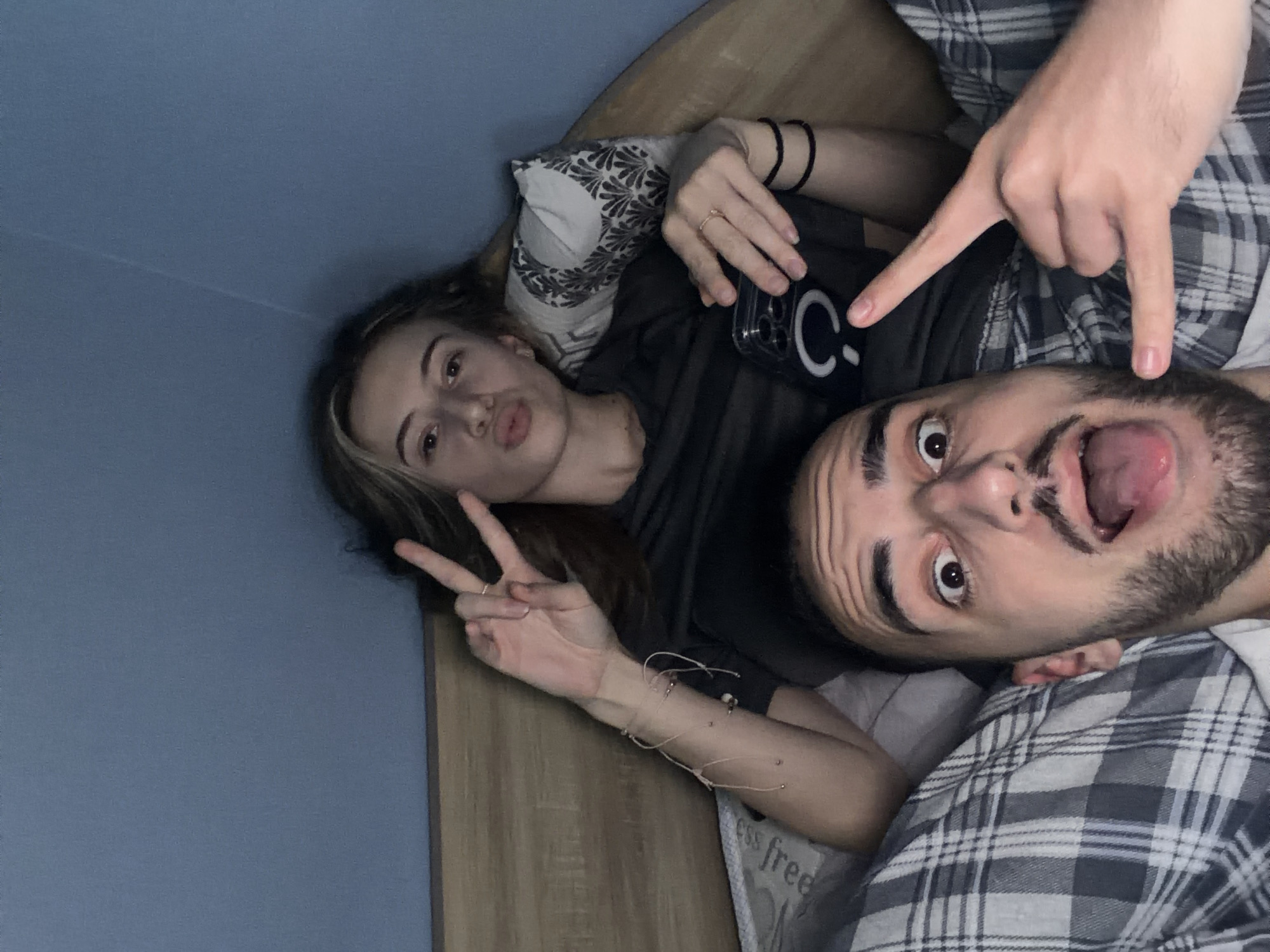 Two individuals are lying in a bed. A woman is on the left side of the frame, making a peace sign with her left hand and holding a phone in her right hand. She has brown hair with blonde highlights. A man is on the right side of the frame, sticking his tongue out, with fingers of another hand pointing down at his face. The room has blue walls. The location is not discernible from the image.