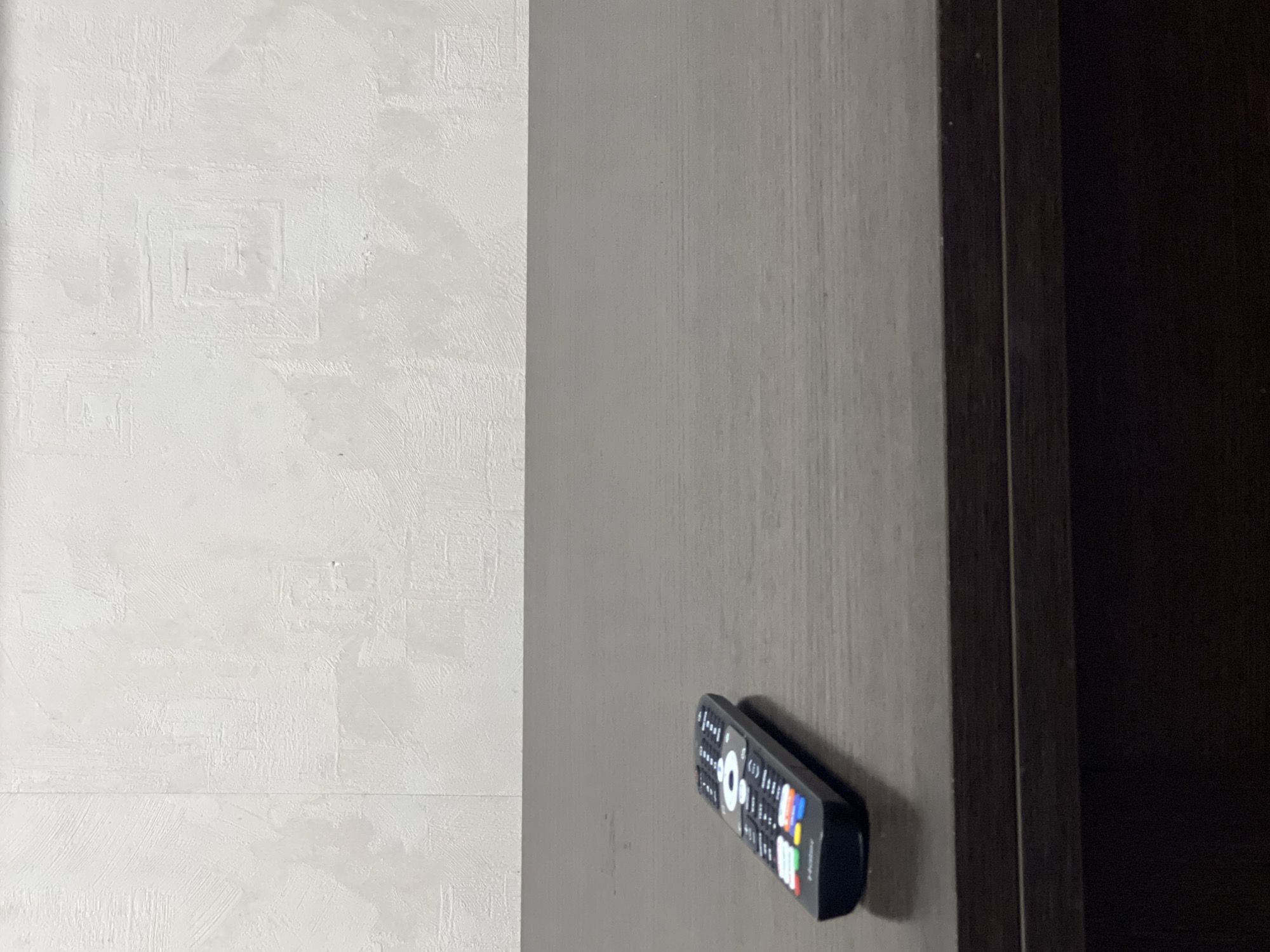 The image depicts a black television remote control resting on a dark brown, wood-grained surface. The surface appears to be a table or shelf, with a lighter-colored, textured wall in the background. The wall has a subtle pattern, possibly wallpaper, with visible brushstrokes or textured lines. The lighting is soft, creating a slight shadow from the remote. The scene is static and devoid of any human presence or activity. There is no information in the image that would allow for its location in Yekaterinburg, Russia.