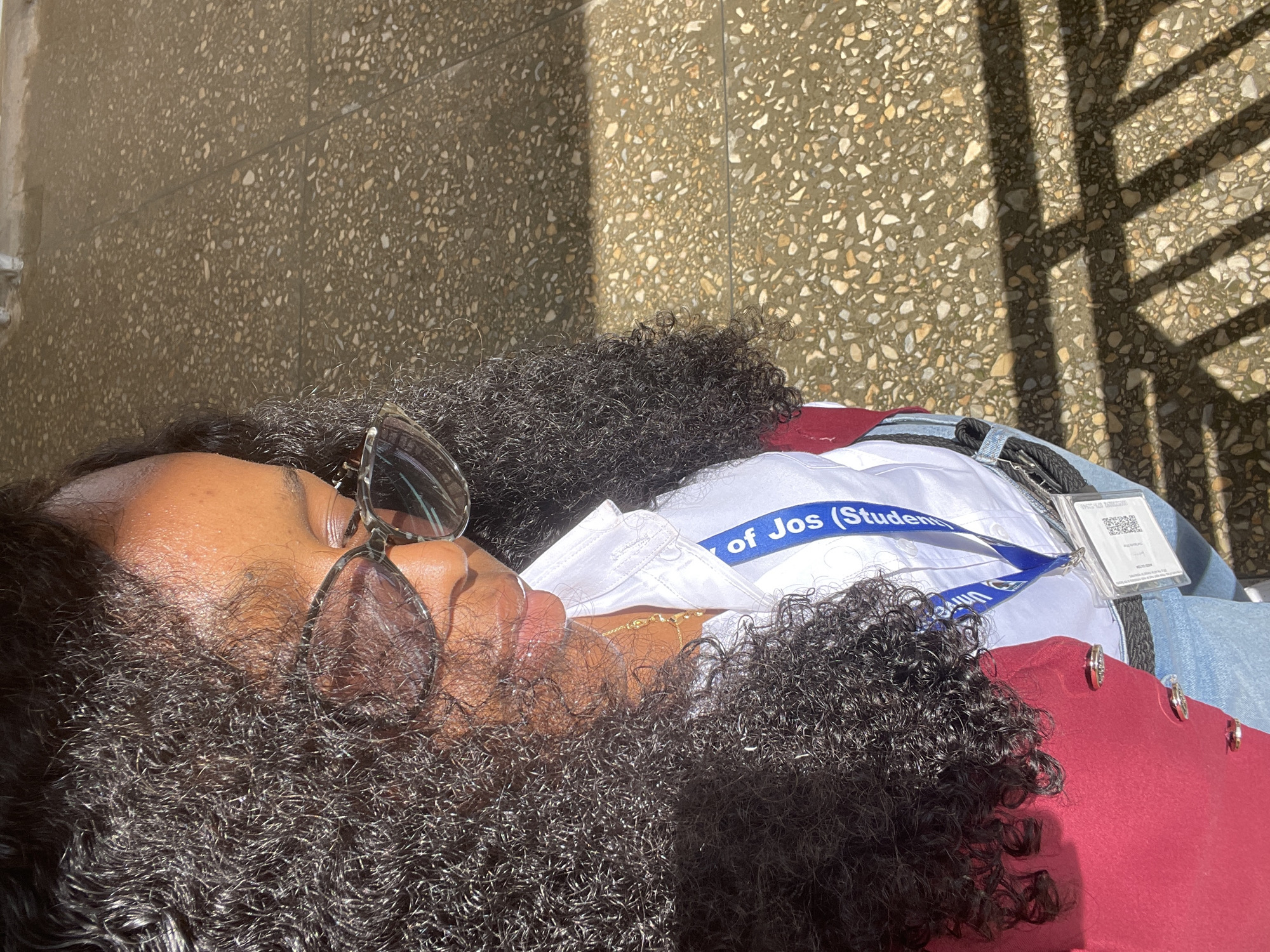 In Rafin Par, Nigeria, a woman with dark, curly hair and clear-framed glasses stands outdoors, illuminated by sunlight casting shadows on the textured ground. She is wearing a white collared shirt, a maroon jacket, and light blue jeans. A blue lanyard with "University of Jos (Student)" printed on it is visible around her neck, attached to an identification card in a clear plastic holder. The ground appears to be paved with small, multi-colored stones.