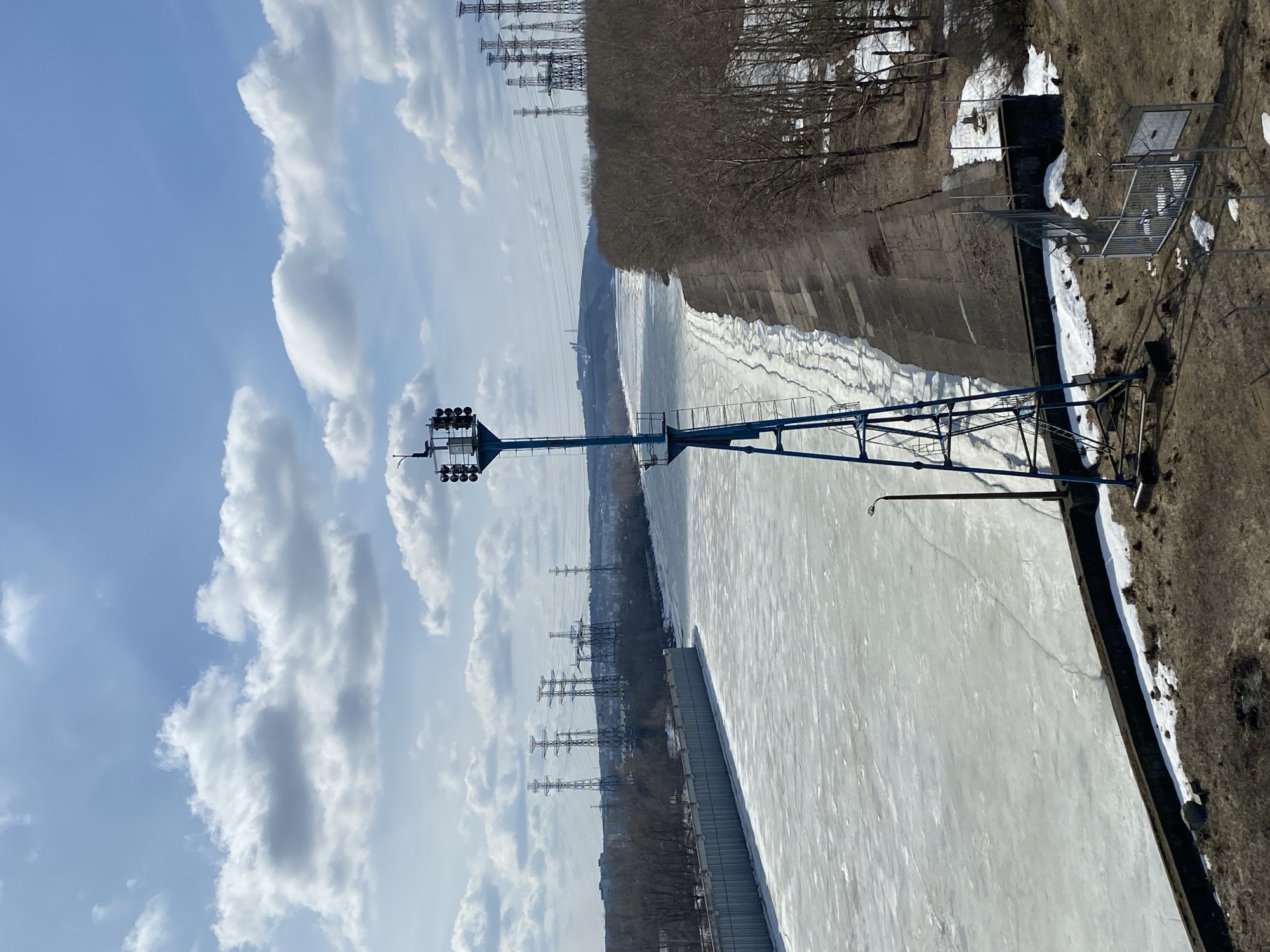 The image shows a partly frozen body of water, likely the Kama River, running through Chaikovsky, Russia. An angled view reveals a concrete embankment on one side of the river, with patches of snow and sparse vegetation along its edge. A blue-painted metal structure with multiple spotlights extends out over the water, possibly a river monitoring station or lighting fixture. Power lines are visible on the opposite shore, suggesting an urban or industrial area. The sky is partly cloudy. The overall scene indicates a cold, possibly late-winter or early-spring day.