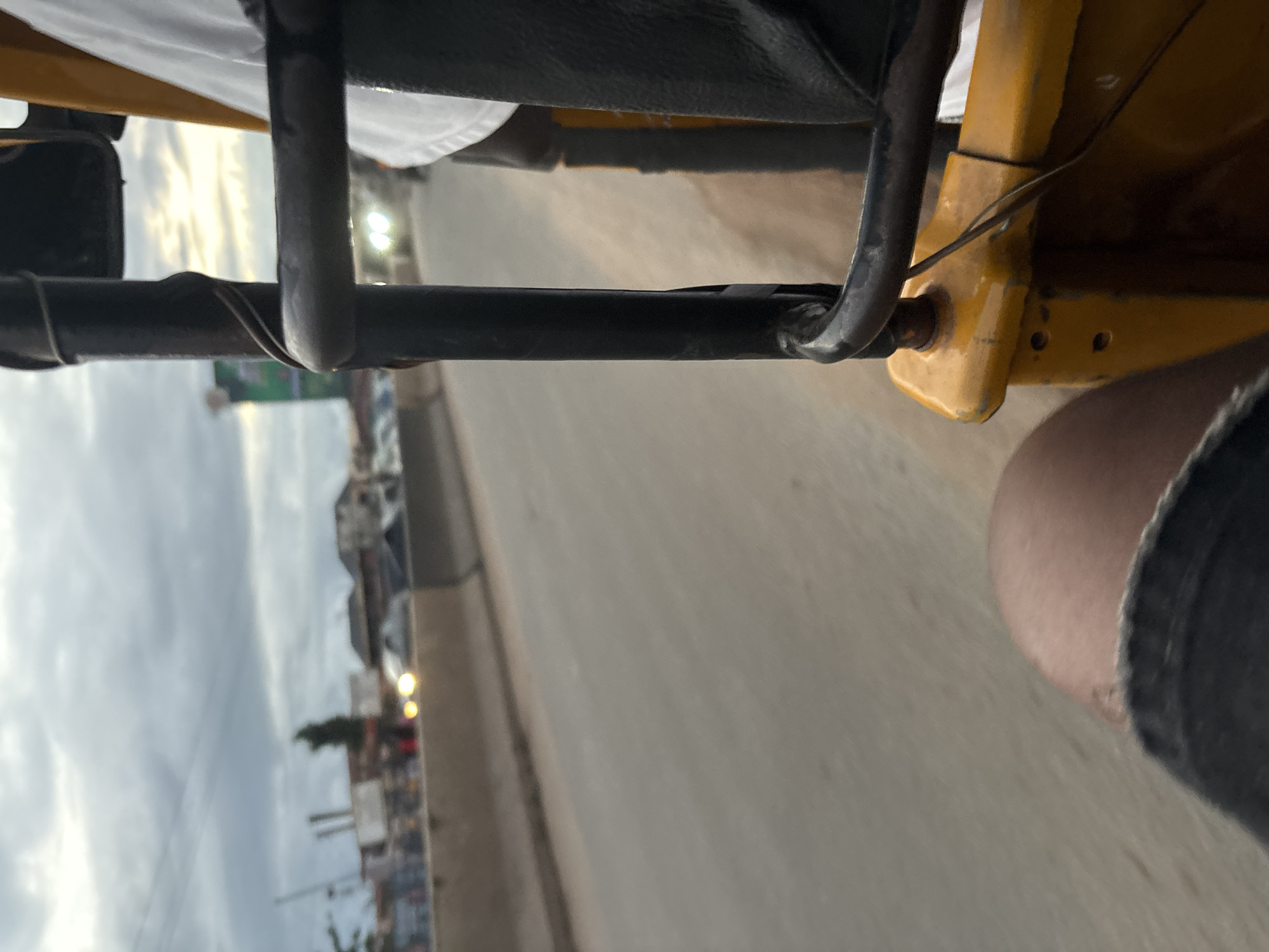 An extreme close-up shot captures the perspective of someone riding in a yellow auto-rickshaw, or Keke Napep, in Onitsha, Nigeria. The foreground is dominated by the rough texture of the auto-rickshaw's yellow frame and a dark metal bar, with a sliver of the rider's thigh visible at the bottom left, clad in dark denim shorts. The background is a blur of motion, indicating the vehicle is traveling. A concrete barrier separates the road from what appears to be a roadside market or urban settlement, with buildings and indistinct figures visible. The sky is overcast with dramatic clouds, suggesting either dawn or dusk. The overall impression is one of movement and the everyday hustle of urban life in Onitsha.