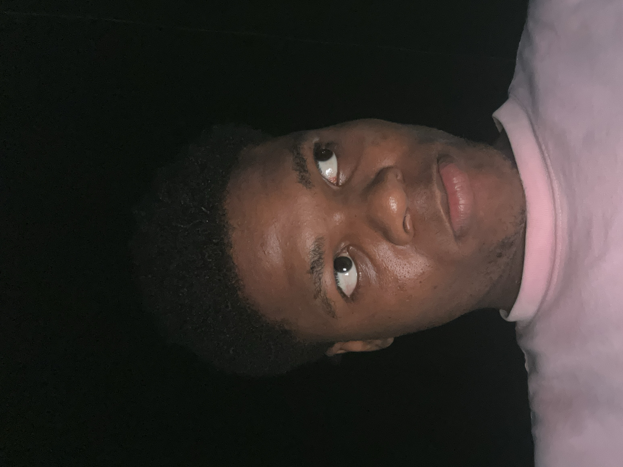 The image is a close-up, head-and-shoulders shot of a young Black male with short, curly dark hair. He is looking to his right with his mouth slightly ajar. He is wearing a light pink collared shirt. The background is entirely black, indicating a dark environment. The lighting is focused on the subject's face, highlighting his skin texture and the sheen of sweat on his forehead and cheeks. The overall impression is that this is a selfie taken in low-light conditions, possibly at night. There is no visible indication of the location being the city of Ihiagwa, Nigeria.