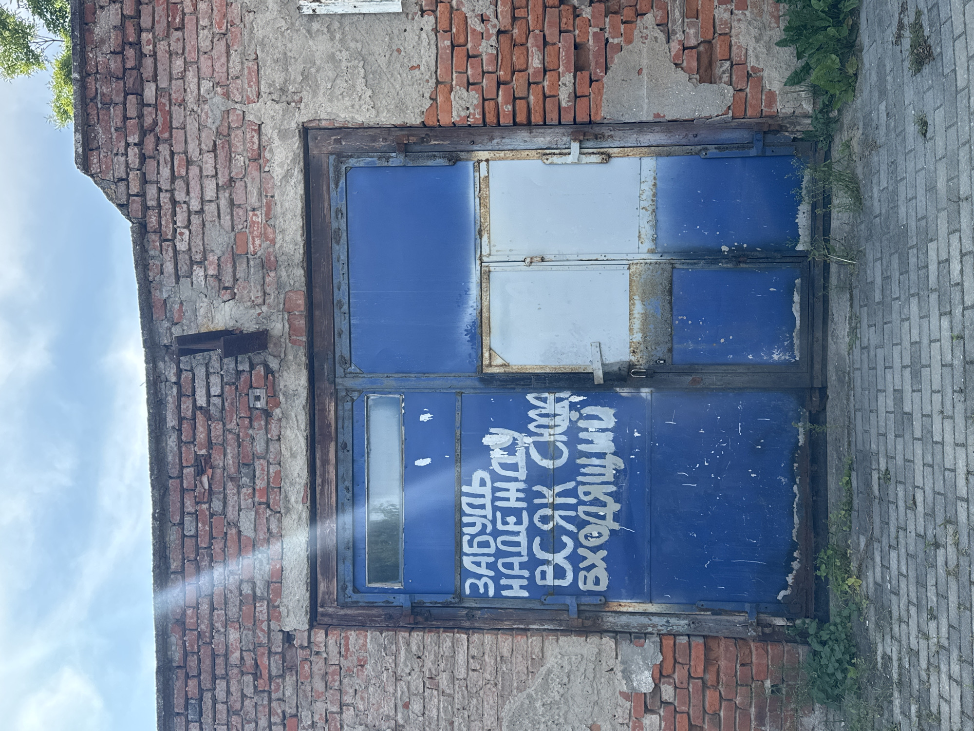 An exterior, eye-level, vertical, medium shot captures a weathered, blue metal garage door set within a dilapidated brick wall under a cloudy sky. The brick wall shows signs of decay with crumbling mortar and exposed brickwork. The garage door itself is divided into two large sections and two smaller ones. White, handwritten text in an unfamiliar script is scrawled across the left-hand section of the large blue door. The right-hand section of the door is a lighter blue, with a smaller, light gray metal door section below it. The door frame and hinges are rusted. Weeds and grass grow around the base of the door and against the brick wall. The ground in front of the garage is paved with gray bricks. The image appears to be taken in Bagrationovsk, Russia, given the context of the text on the door which reads "Забудь надежду всяк сюда входящий" which translates to "Abandon all hope, ye who enter here". This phrase is a famous quote from Dante Alighieri's Inferno, often used to convey a sense of despair or ultimate finality.