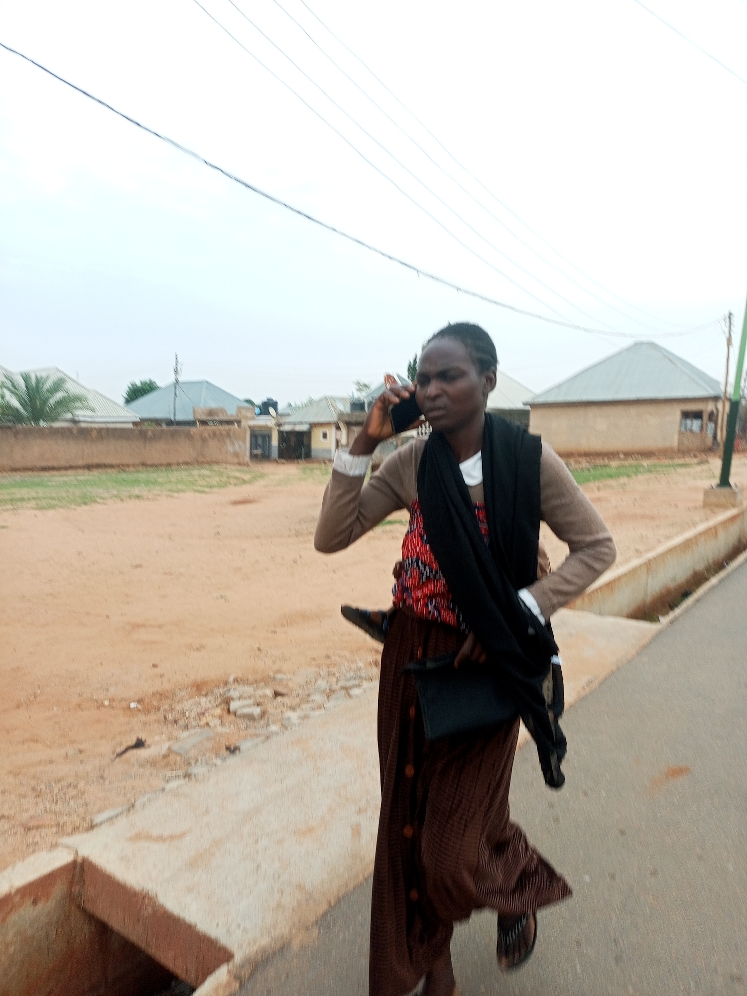 An adult woman is walking along a paved road, talking on a mobile phone. She is wearing a brown cardigan, a red and black patterned top, a long brown button-down skirt, a black scarf draped around her neck, and sandals. She is carrying a black clutch bag. The road has a concrete curb and a drainage ditch. Beyond the road, there is a sandy area with scattered debris and buildings, including houses with corrugated metal roofs. Utility poles and overhead wires are visible in the sky. The scene appears to be in a residential area of Tempure, Nigeria.