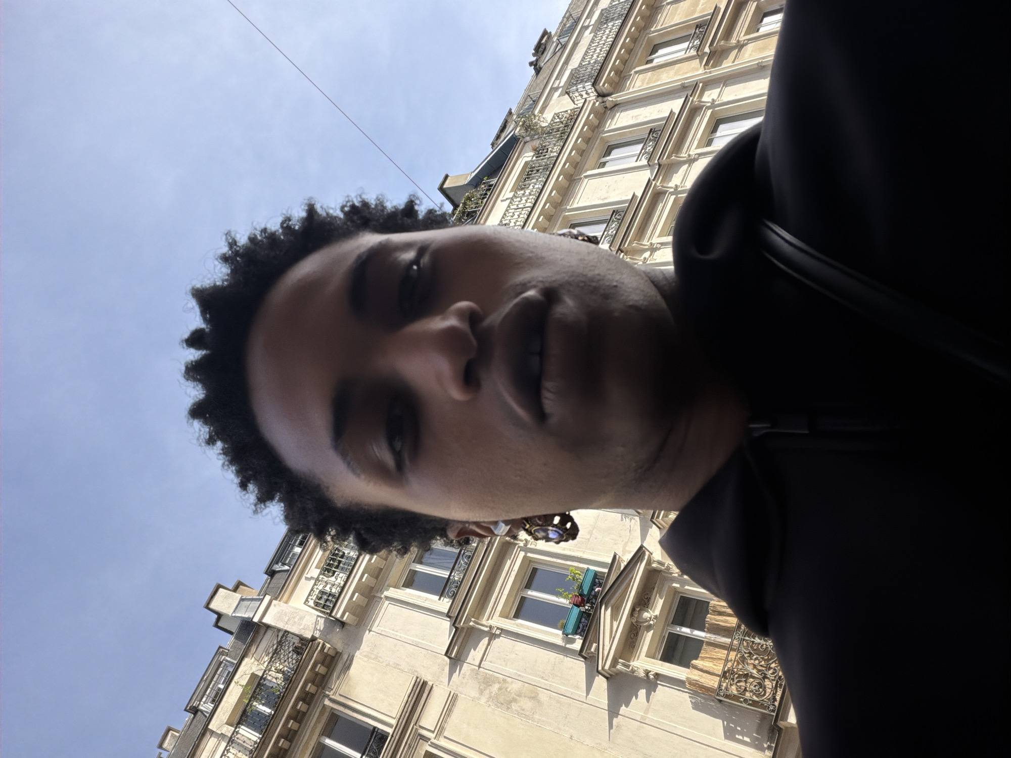 A close-up, low-angle selfie features a person looking upwards, their face partially visible from the chin to just above the eyes, with dark, curly hair framing their head. They are wearing a dark garment, possibly a hoodie or jacket.
The background reveals a tall, light-colored building consistent with Parisian architecture, featuring multiple floors with ornate wrought-iron balconies and tall windows. Some windows have decorative elements or dark awnings, and a few balconies display small planters with greenery. The building's details are highlighted by bright sunlight, suggesting a clear day. Above the building, a vast expanse of clear blue sky is visible, with a faint, thin line (possibly a wire or cable) traversing the upper left portion of the frame. The overall impression is one of sunny weather in an urban European setting, specifically Paris, France. No visible text is present in the image.