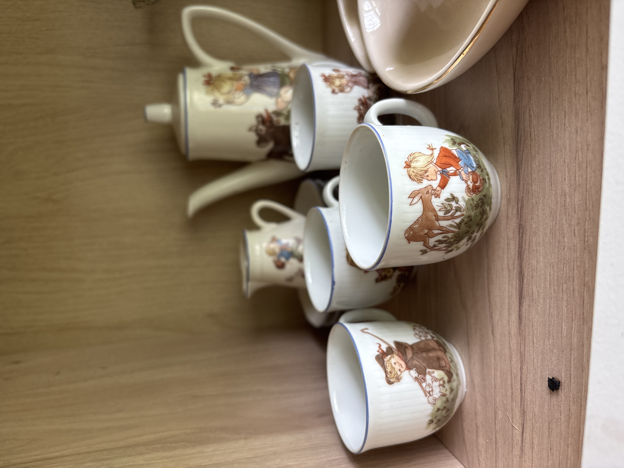 In Borki, Russia, a wooden shelf displays a collection of ceramic tea set items. The primary subjects are four white ceramic cups, a matching teapot, and a small creamer. Each piece is adorned with hand-painted illustrations featuring children interacting with animals, specifically a girl with sheep, a girl feeding a deer, and a girl with a bird. The cups and teapot have blue trim around their rims. In the background, a large teapot with a lid and handle is visible. To the right of the cups, a pink ceramic dish with a gold rim is partially in view. The shelf itself is made of light-colored wood with a visible grain pattern. A small, dark, irregularly shaped object, possibly a piece of lint or debris, rests on the shelf in the foreground. The lighting suggests an indoor setting, likely a cabinet or display case, with the light source coming from the front.